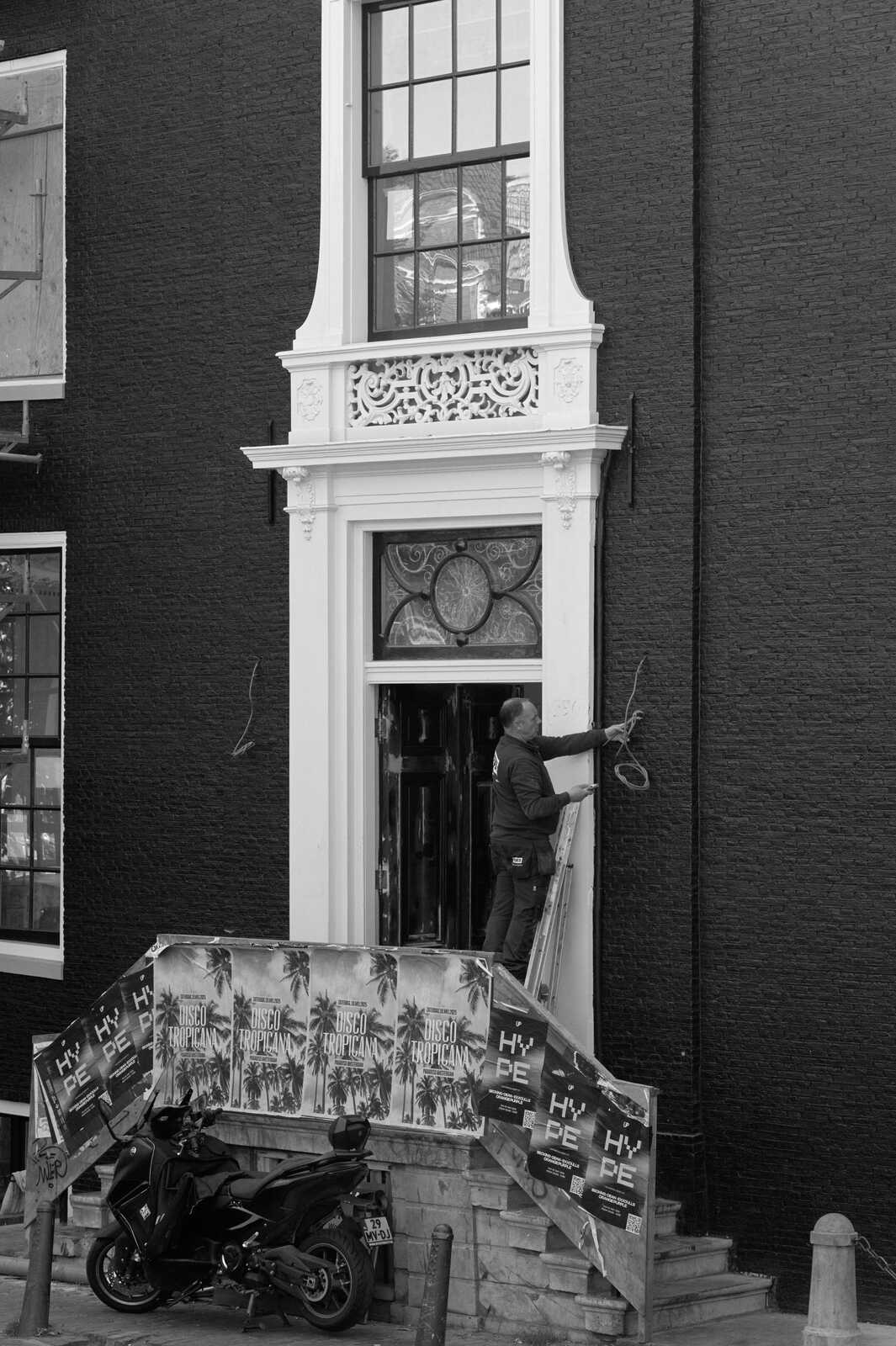 A man in a dark jacket is carrying a panel up a flight of stairs in front of a black building with ornate window trim. A scooter is parked below. Posters are visible.