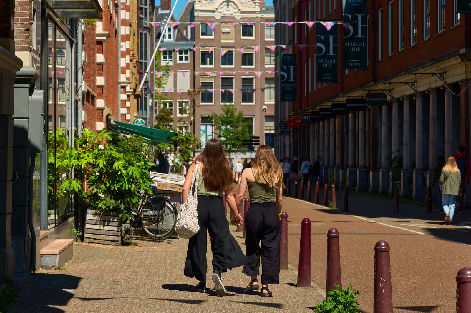 Two women walk hand-in-hand down a cobblestone street lined with shops and red bollards. A bicycle leans against a building. Bright daylight and greenery are visible.