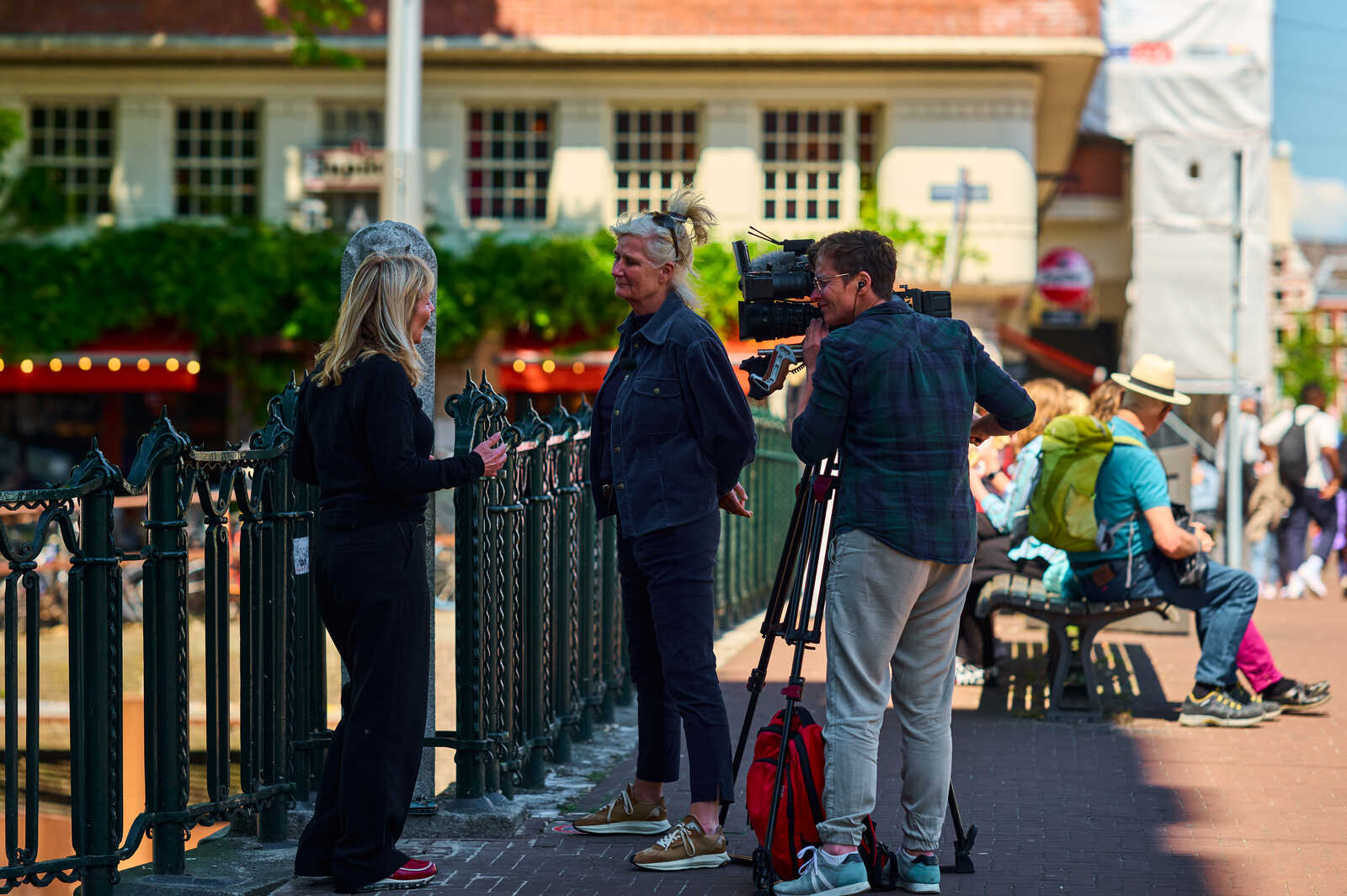 Two women converse near an iron railing. A man operates a camera on a tripod, positioned on a paved walkway with a building and greenery in the background. A person wearing a hat sits nearby.