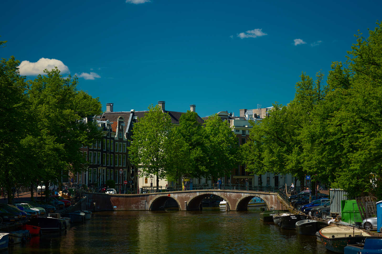 A canal with a stone bridge, lined with trees and colorful buildings. Boats and cars are parked along the waterway. A person is walking on the bridge. Blue sky above.
