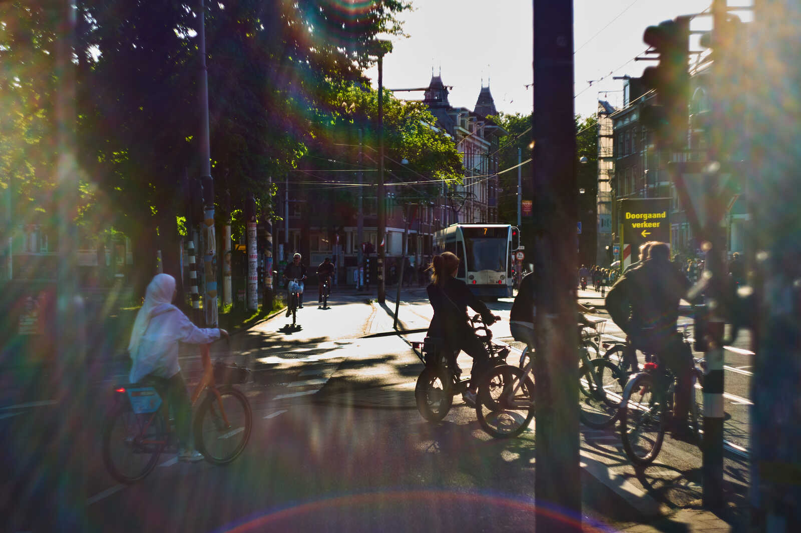 A street scene with people on bicycles and a bus in sunlight. Buildings and trees line the sides of the street. A sign reads Deorsaand verkeer.