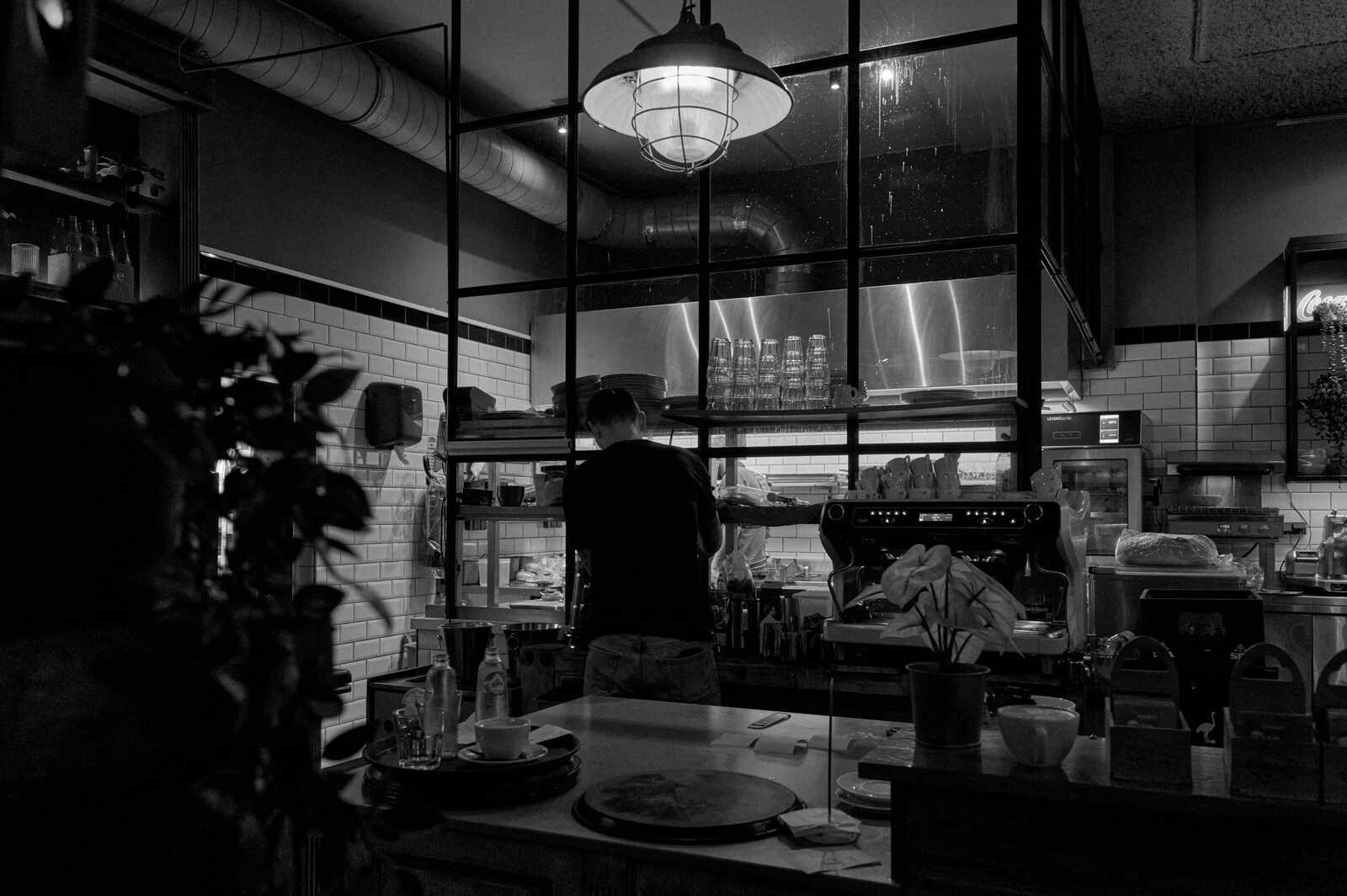 Black and white photo of a cafe interior. A barista stands behind a counter with a coffee machine. There are dishes and cups on the counter and shelves. A large window shows a busy kitchen area.