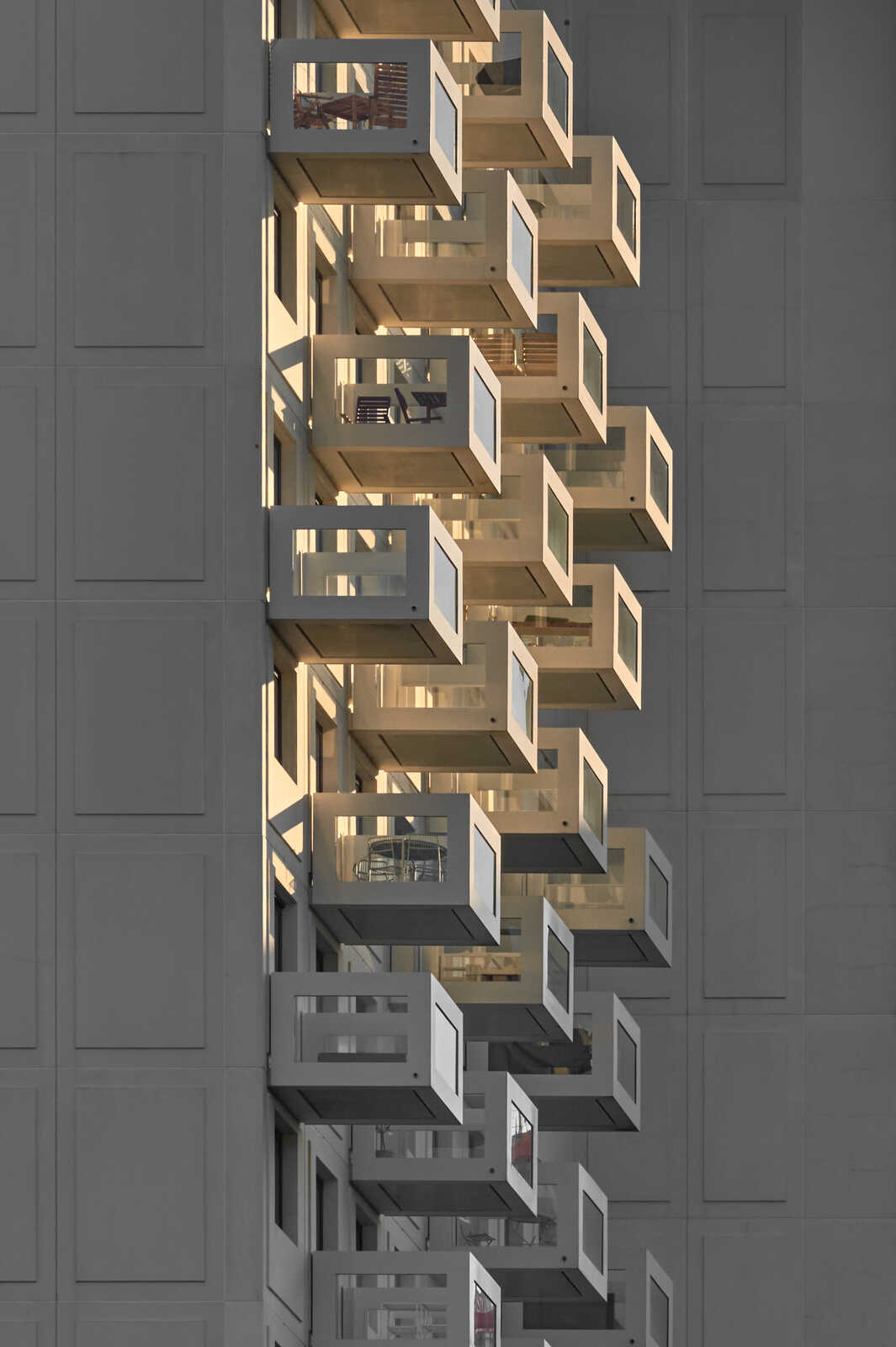 A vertical view of several stacked balconies with open, rectangular railings and windows. The balconies appear to be part of a modern building with a gray facade. Bright light illuminates the balconies.