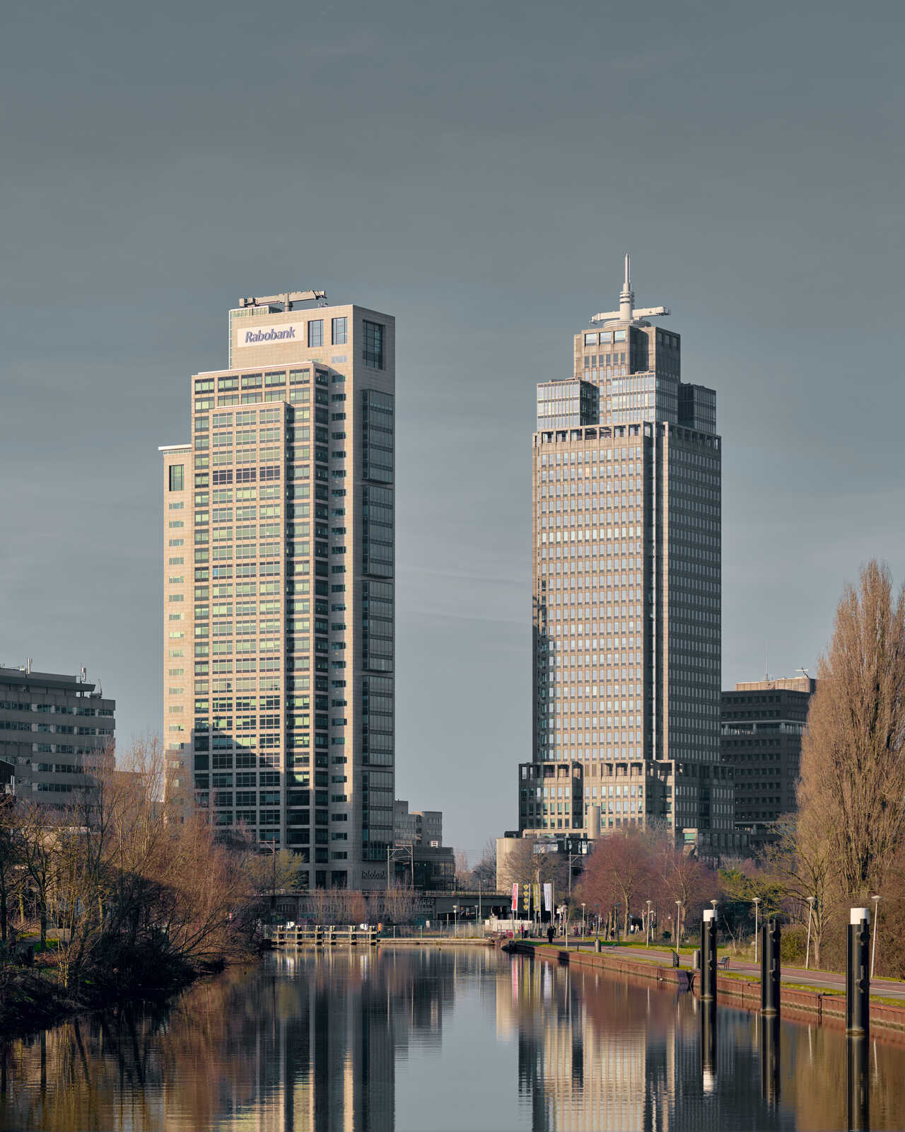 Tall, modern office building with a spire and "Rabobank" signage. Reflects in a calm river, with trees and other buildings in the background. Sky is overcast.