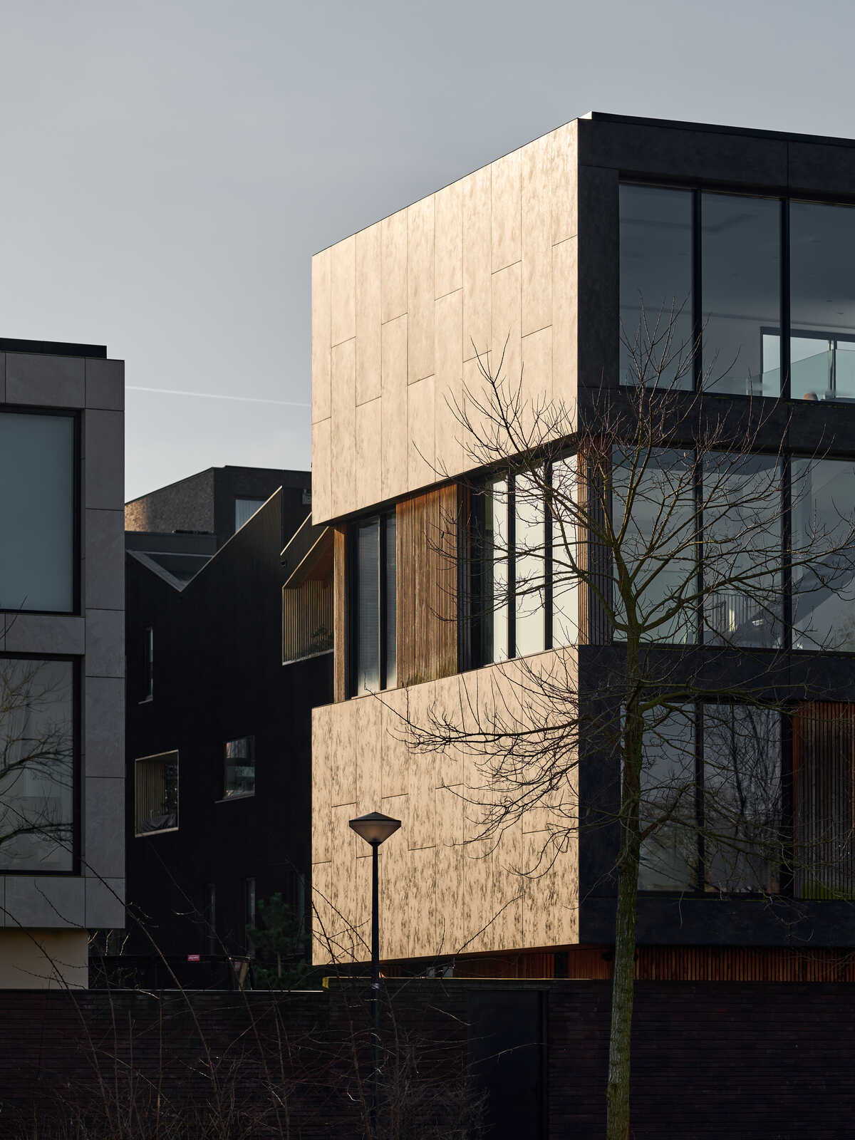Modern building with wood-paneled facade and large windows. Dark roofline and surrounding vegetation. Street lamp visible. Black fences and other buildings in background.