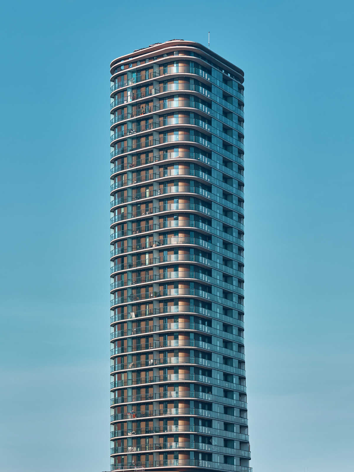 Tall, modern apartment building with numerous balconies. The building has a rectangular shape and is against a clear, blue sky. Rows of windows are visible on each level.