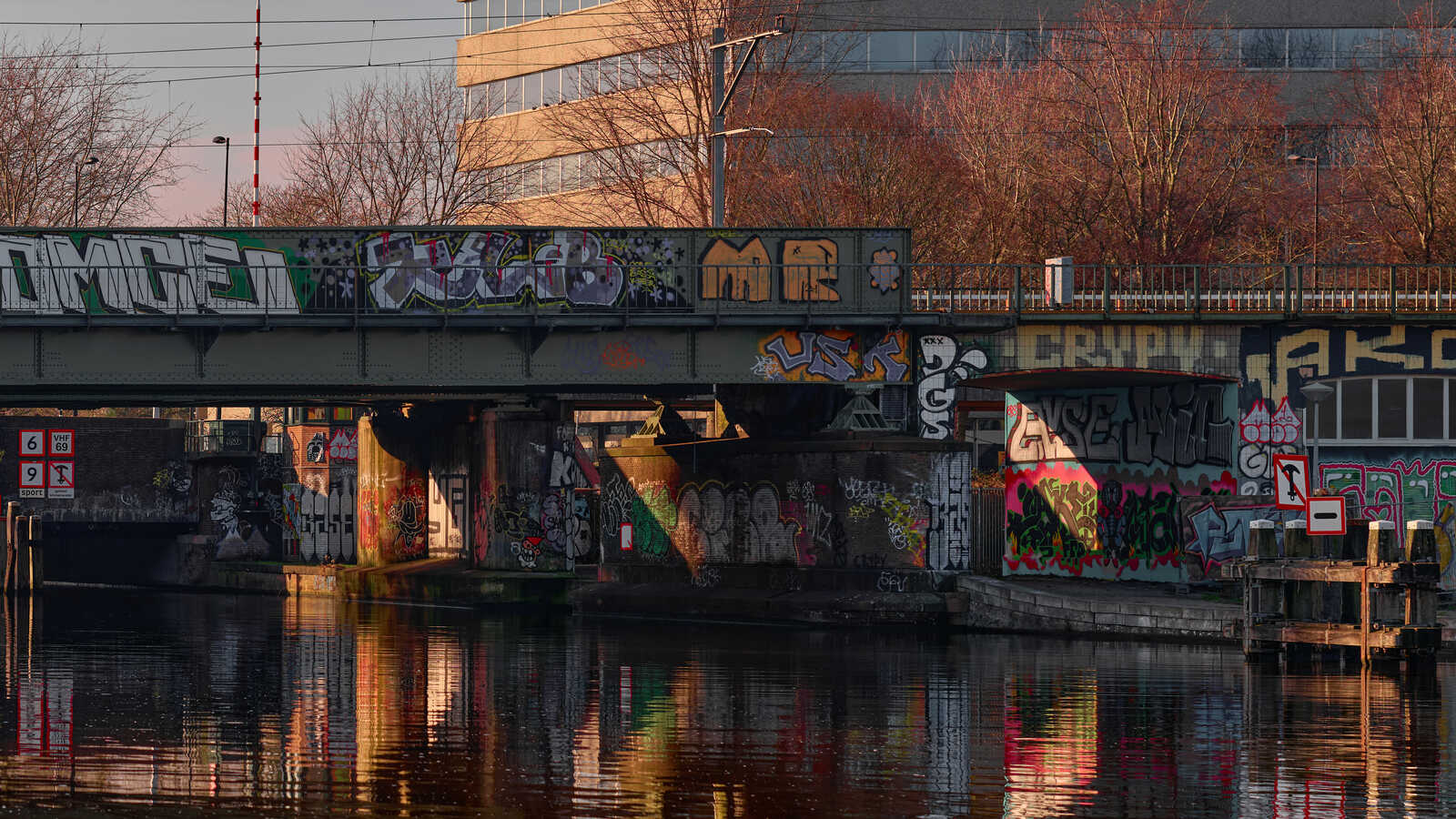 A canal with a graffiti-covered bridge overhead. The water reflects the vibrant colors of the street art. A building is visible in the background.