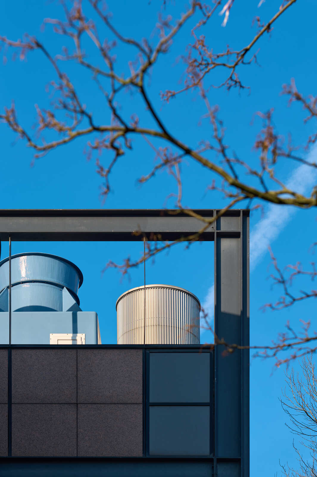 A tall, cylindrical industrial vent with a ribbed exterior sits against a bright blue sky. A grey building with large windows frames the vent. Bare branches are visible in the top left corner.
