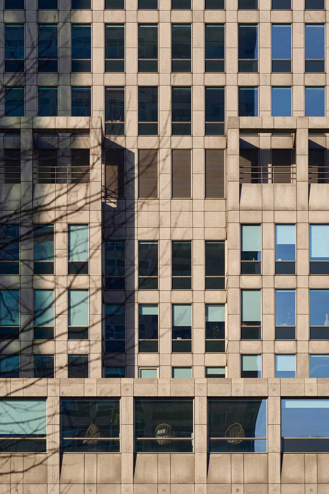 A facade of a tall building. It features a grid of square windows with dark frames and reflective glass. The building’s exterior is light beige with horizontal lines.