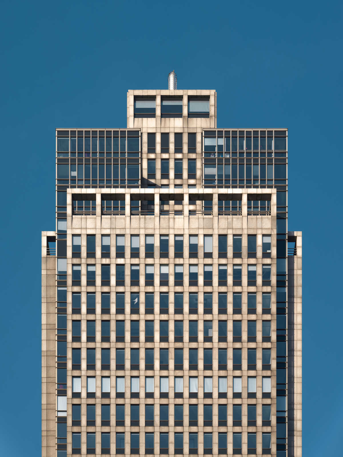 Tall, beige building with a grid of square windows and a flat roof. The sky is a solid, bright blue in the background. There's a small antenna on top.