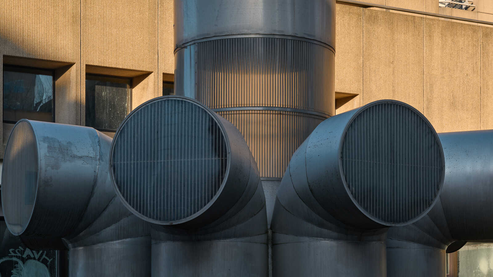 Three large, silver, cylindrical vents with vertical ribs are mounted on a textured, beige wall. A window is visible behind the vents. A small, dark shape is near the bottom left corner.