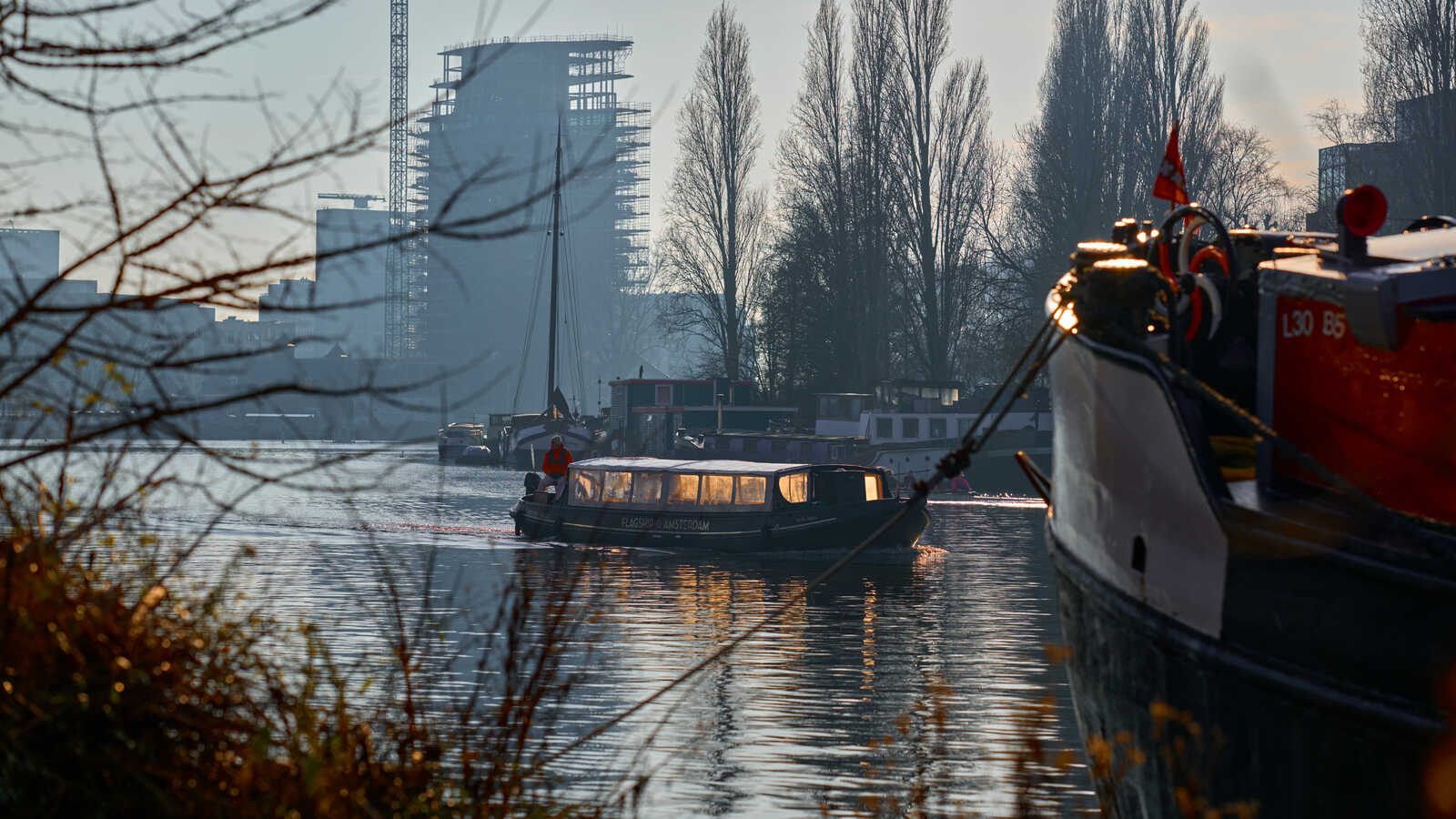 A canal scene with a houseboat, a narrowboat, and a partially constructed building in the background. Water reflects the sky and boats. Branches frame the left side. A mast is visible.