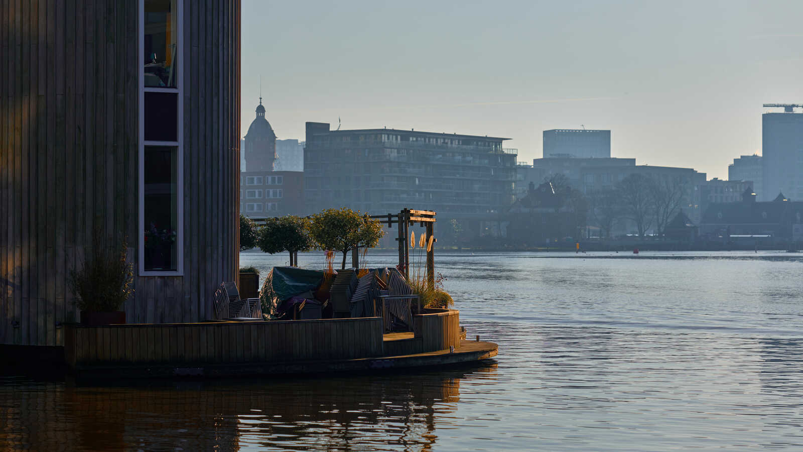A wooden houseboat with outdoor seating and potted plants floats on a calm waterway. City buildings are visible in the hazy distance. A blurred flag is visible on the boat.