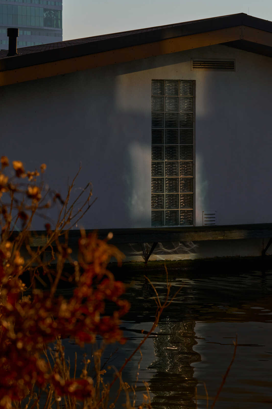 A modern building with a grid window reflecting light on a calm body of water. Bare branches are in the foreground. The sky is a gradient of gray and orange.