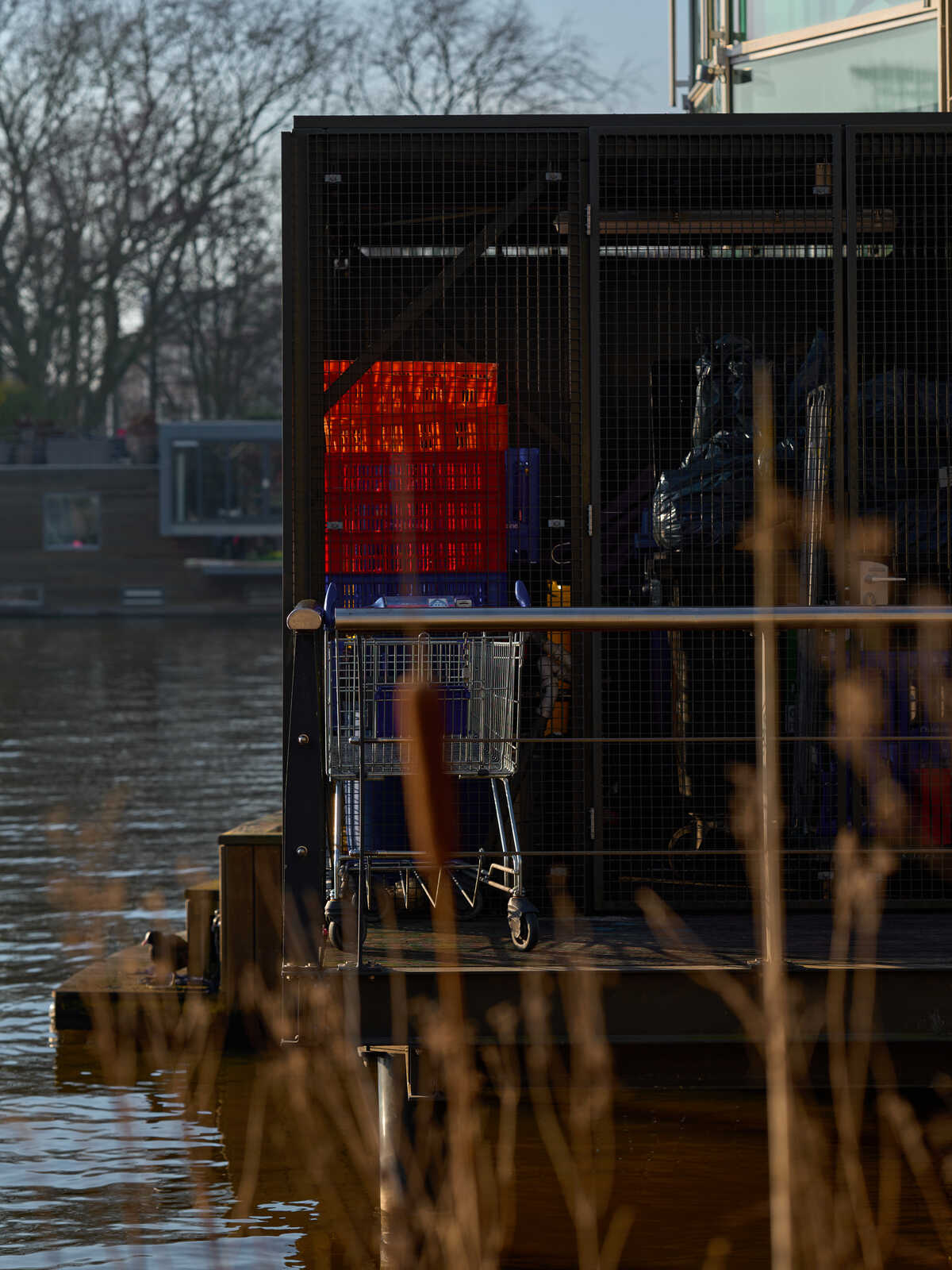 A shopping cart and a stack of red crates rest on a wooden platform near a body of water. A dark metal grate and reeds are in the foreground. A building is visible across the water.