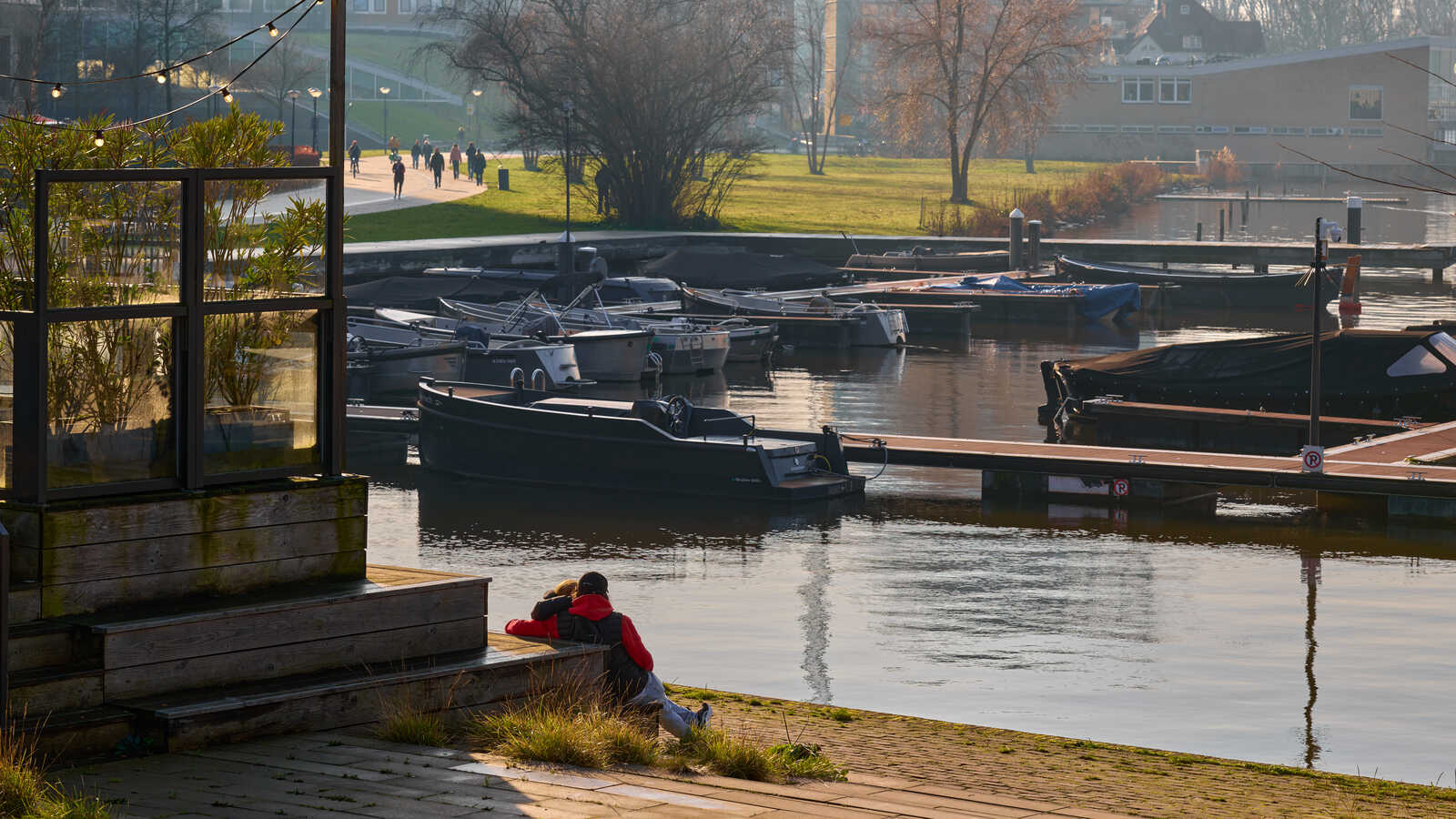 Two people sit on a wooden platform overlooking a canal. Several boats are docked alongside the water. Trees and buildings are visible in the background. The sky is blue.