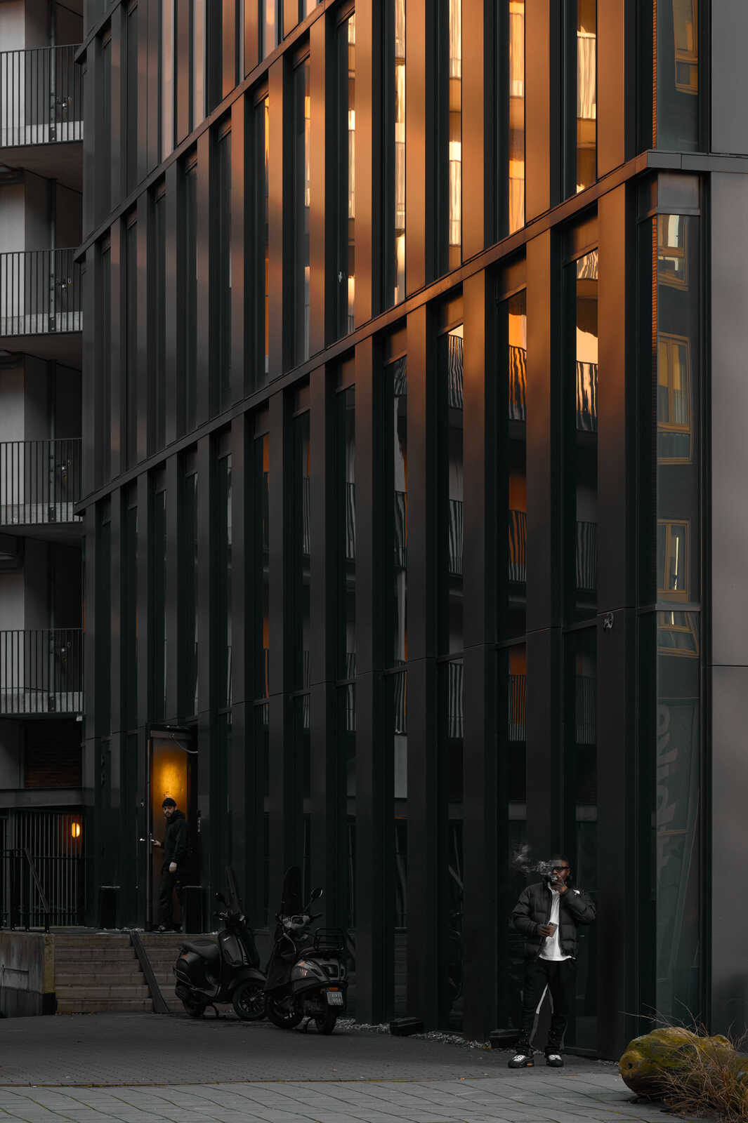 A modern building with dark vertical slats, a scooter, and a person in a puffer jacket stand on a gray concrete walkway. A glass door is visible. Yellow light emanates from within.