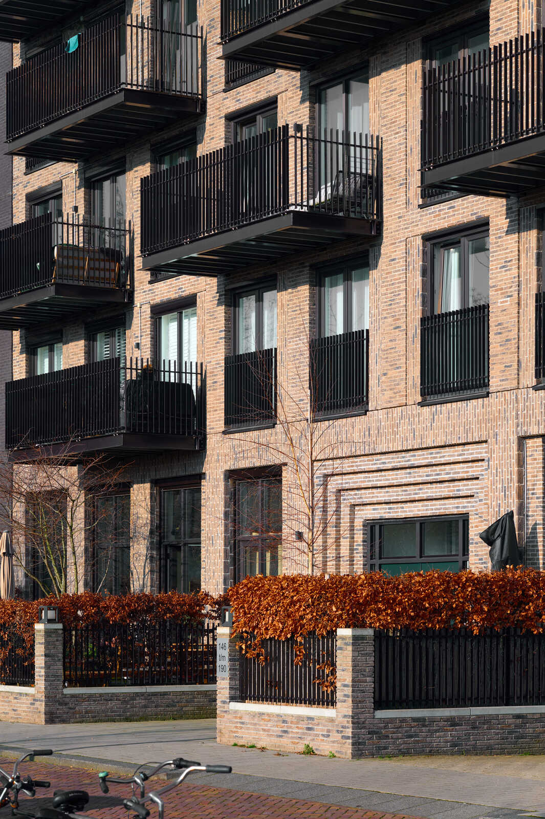 Modern brick apartment building with black wrought iron balconies and a metal fence. Some bicycles are parked on a paved area below. A tree with orange leaves is visible.
