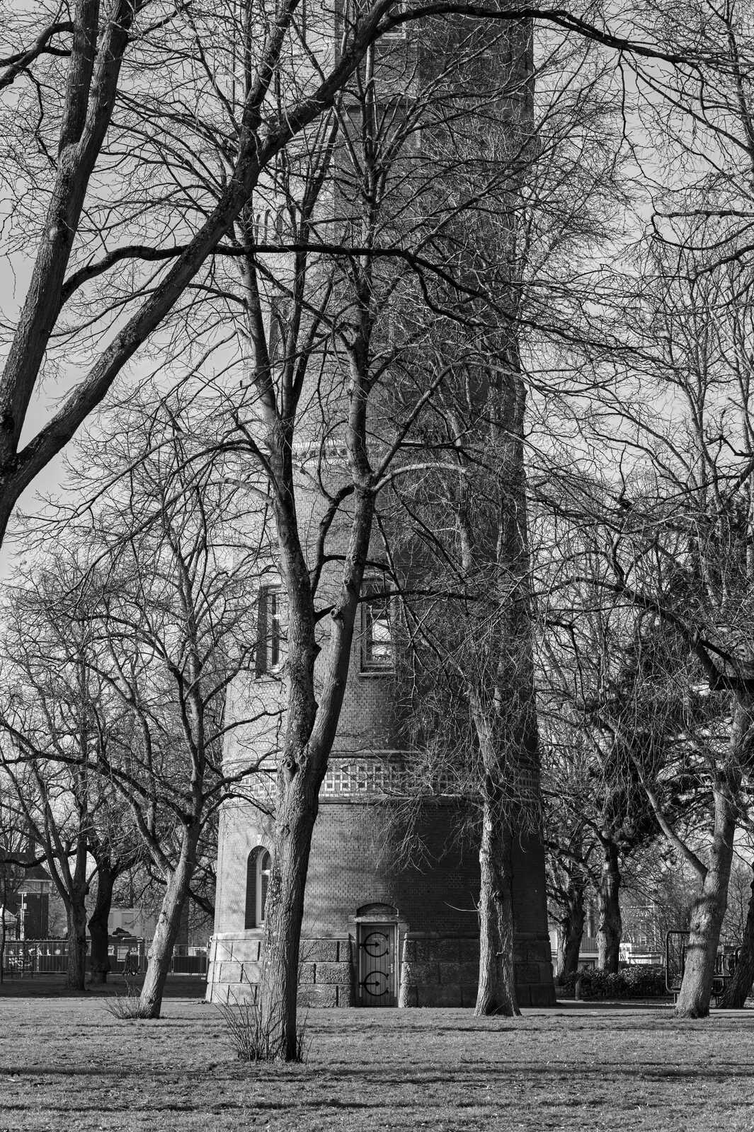 A grayscale photo shows a brick building with arched windows and a tower. Leafless trees frame the structure, with grass and a fence visible in the foreground. Sky is seen above.