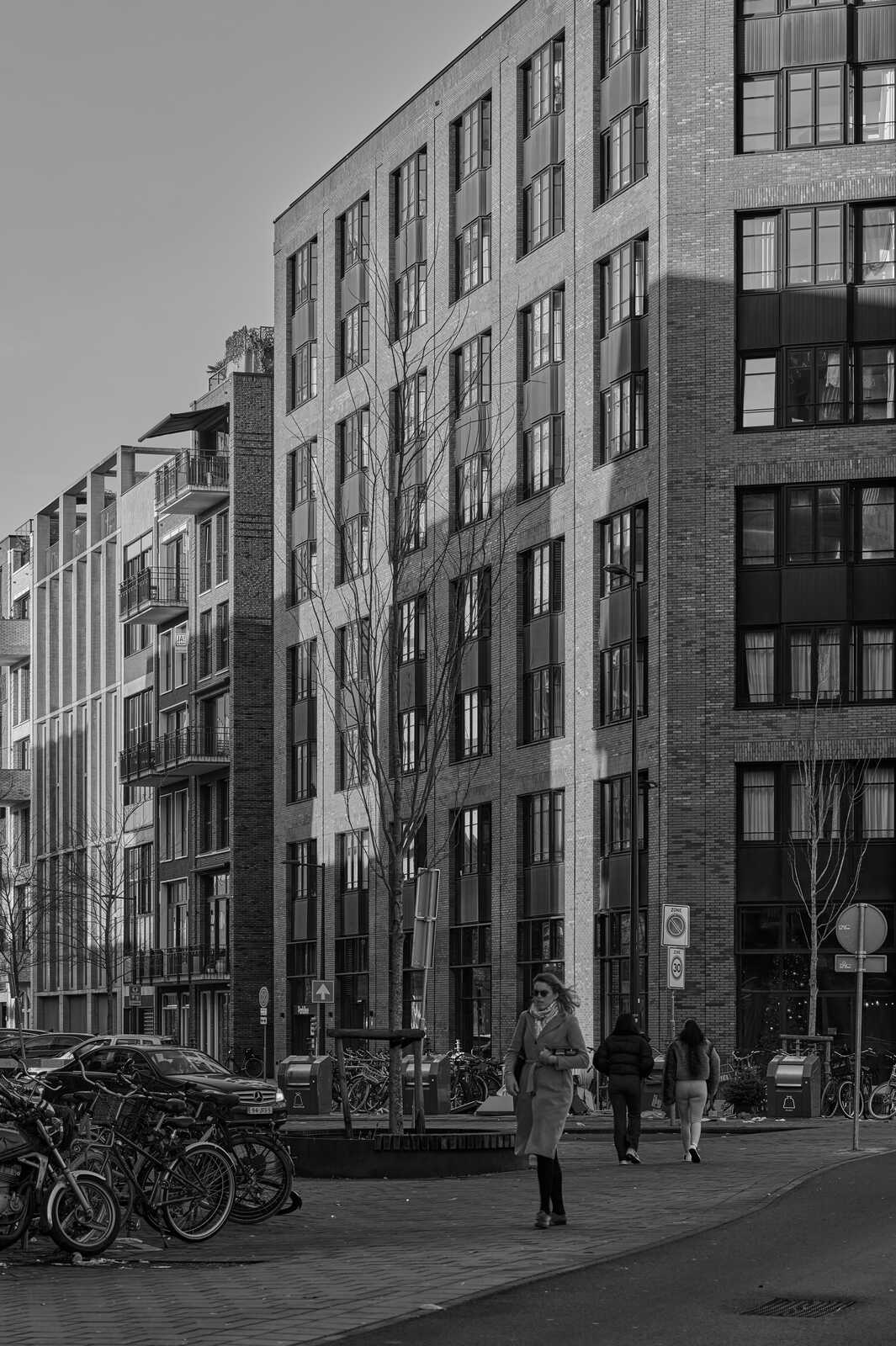 A black and white image showcases a modern brick building with multiple windows and a pedestrian sidewalk. Three individuals are walking along the sidewalk. Bicycles are parked nearby. Signs and a tree are also visible.