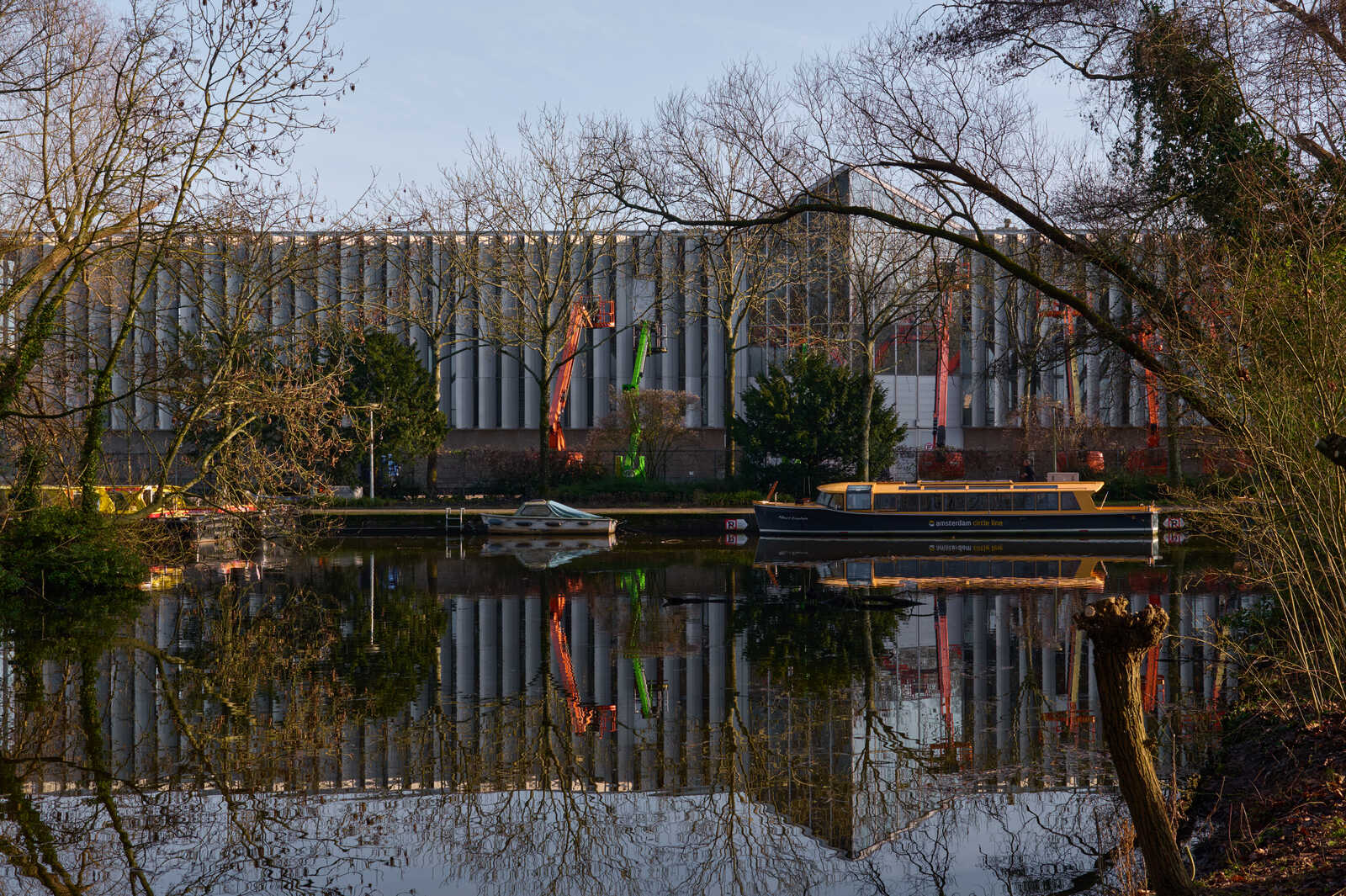 A canal with a boat docked. Red and green flags reflect in the water. Behind the canal is a tall brick building and bare trees. The sky is grey.