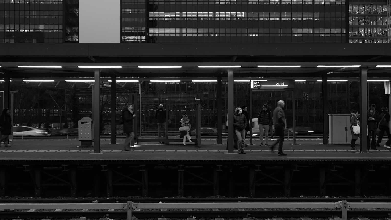 Black and white photo of people on a platform, a train passing, and a building in the background with a sign that reads "Zuid". Several figures are walking. A lamppost stands nearby.