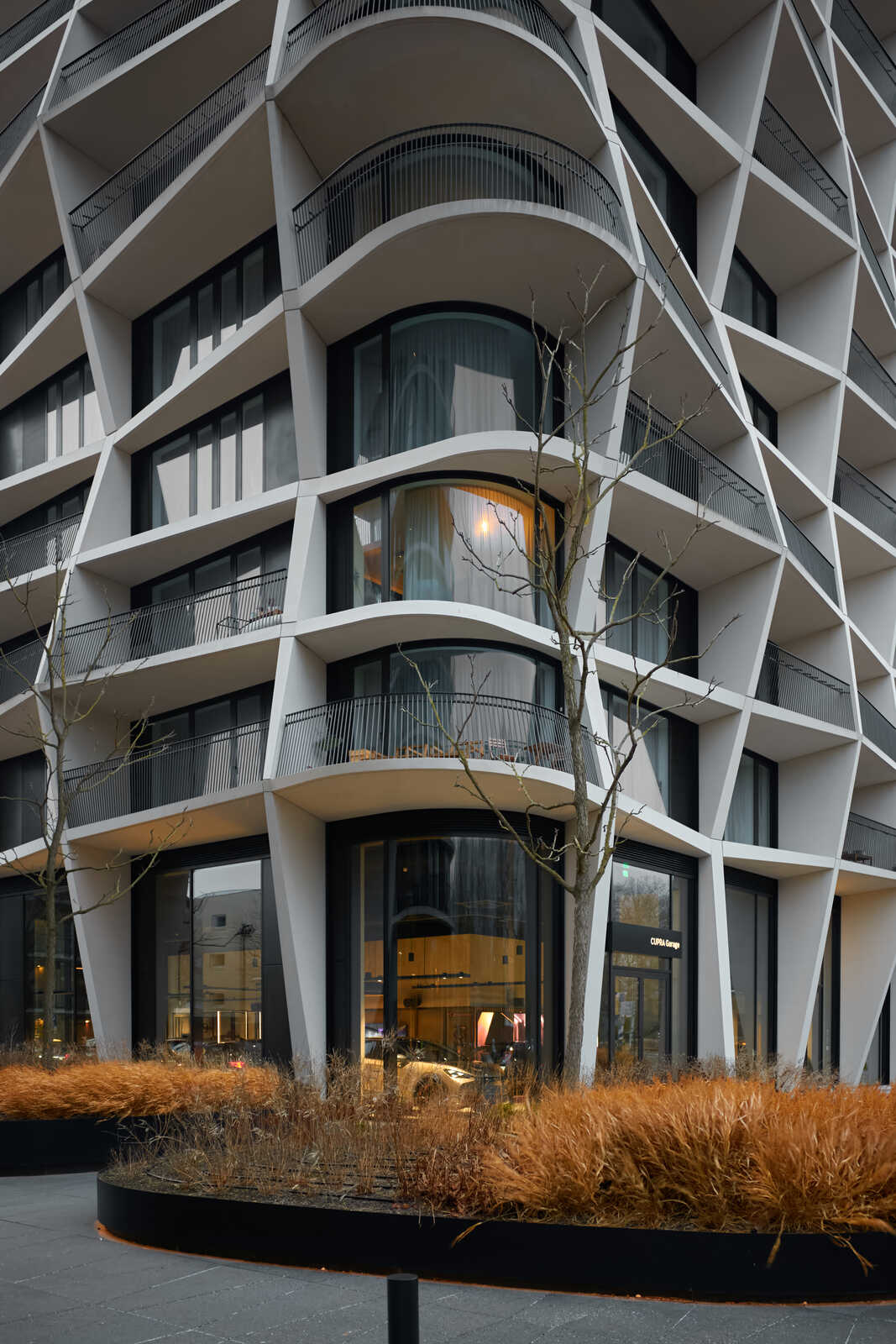 A modern, white building with rounded balconies and large windows. A bare tree and dry grasses are in front of the building. A storefront is visible on the ground level.