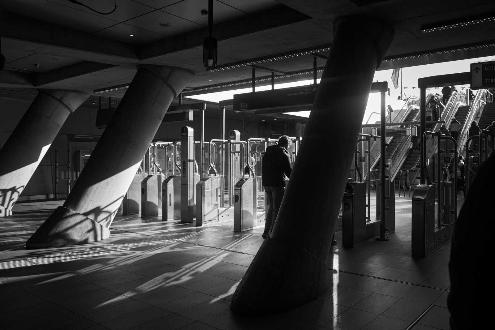 Black and white photo of a ticket gate area. Concrete pillars and overhead signage dominate. A person stands near a gate, bathed in light and shadow. Escalators are visible in the background.