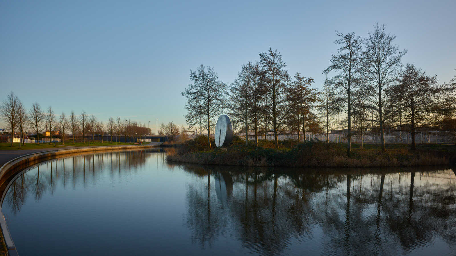 A calm lake reflects bare trees & a spherical sculpture. A paved pathway curves along the water's edge under a dusky sky. Low vegetation lines the bank. The background shows buildings.
