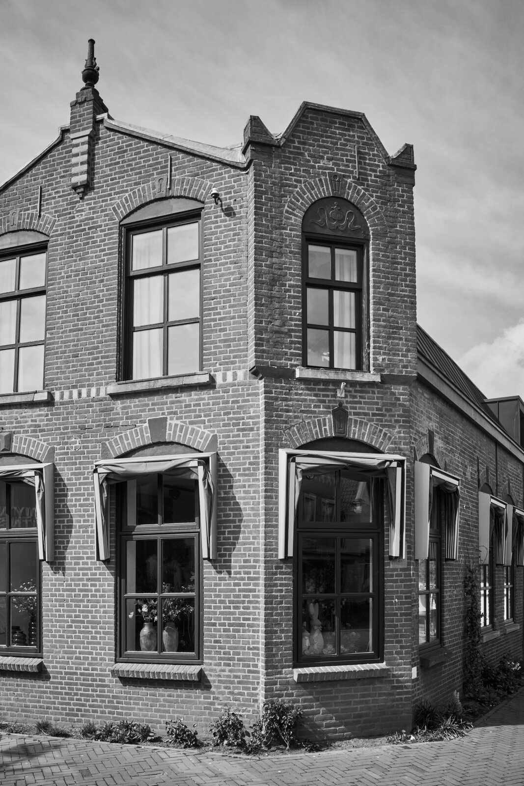 Brick building with a gabled facade. Three tall windows with dark frames and white shutters. A small decorative turret tops the corner. A doorway is visible on the right. Sidewalk in foreground.