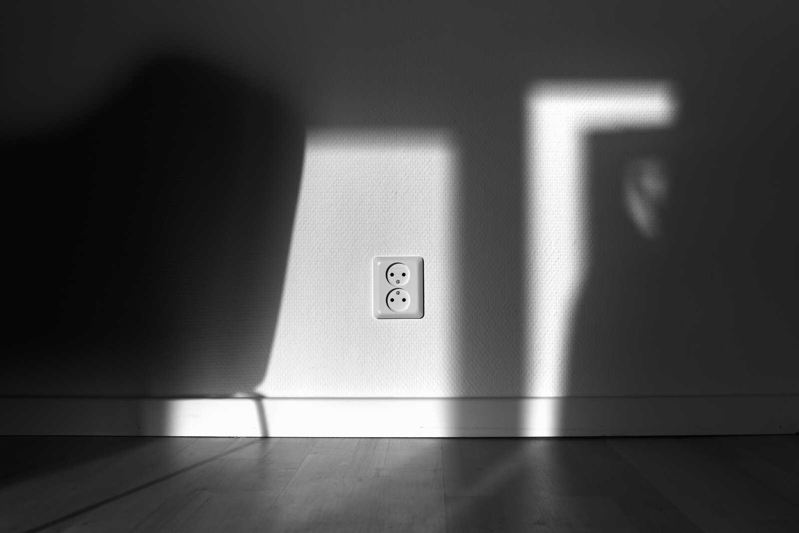 A grayscale photo shows a wall with a white electrical outlet and a long, angled shadow extending across the wall and floor. The floor is wooden.