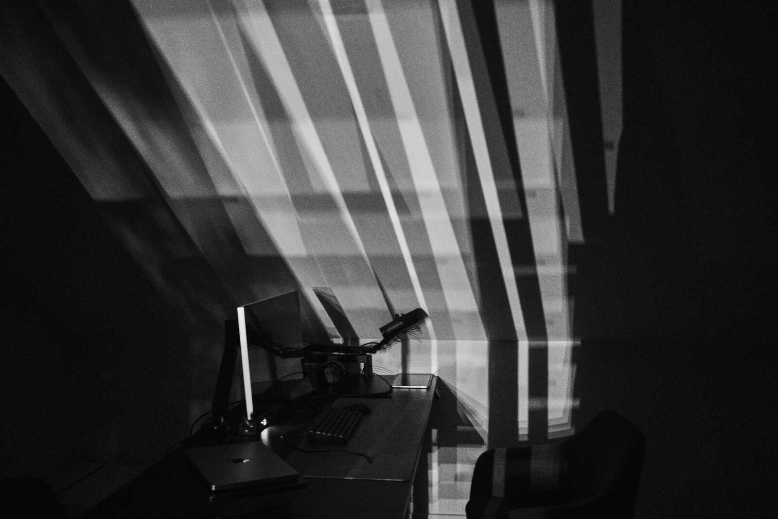 Black and white image of a desk with a monitor, keyboard, and microphone. Long, dark shadows stretch across the surface and wall behind. A laptop is partially visible.