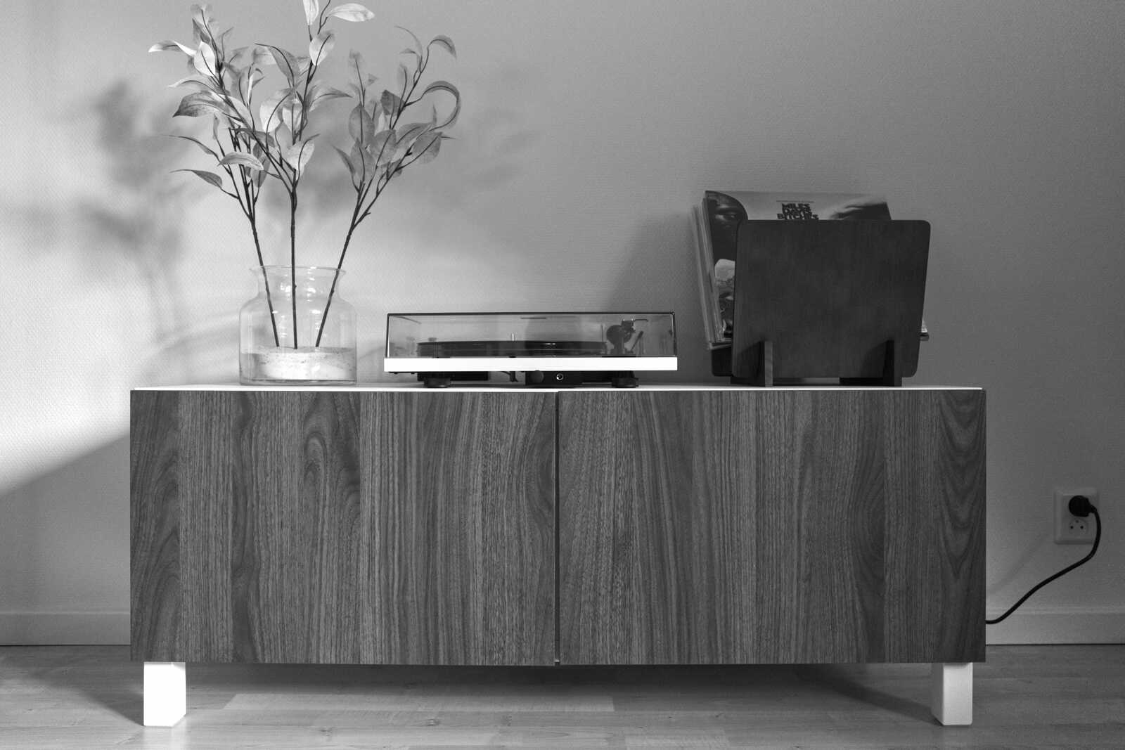 A turntable sits atop a wooden cabinet with two doors and white legs. A vase with green plants rests to the left. A black cord extends from the cabinet.