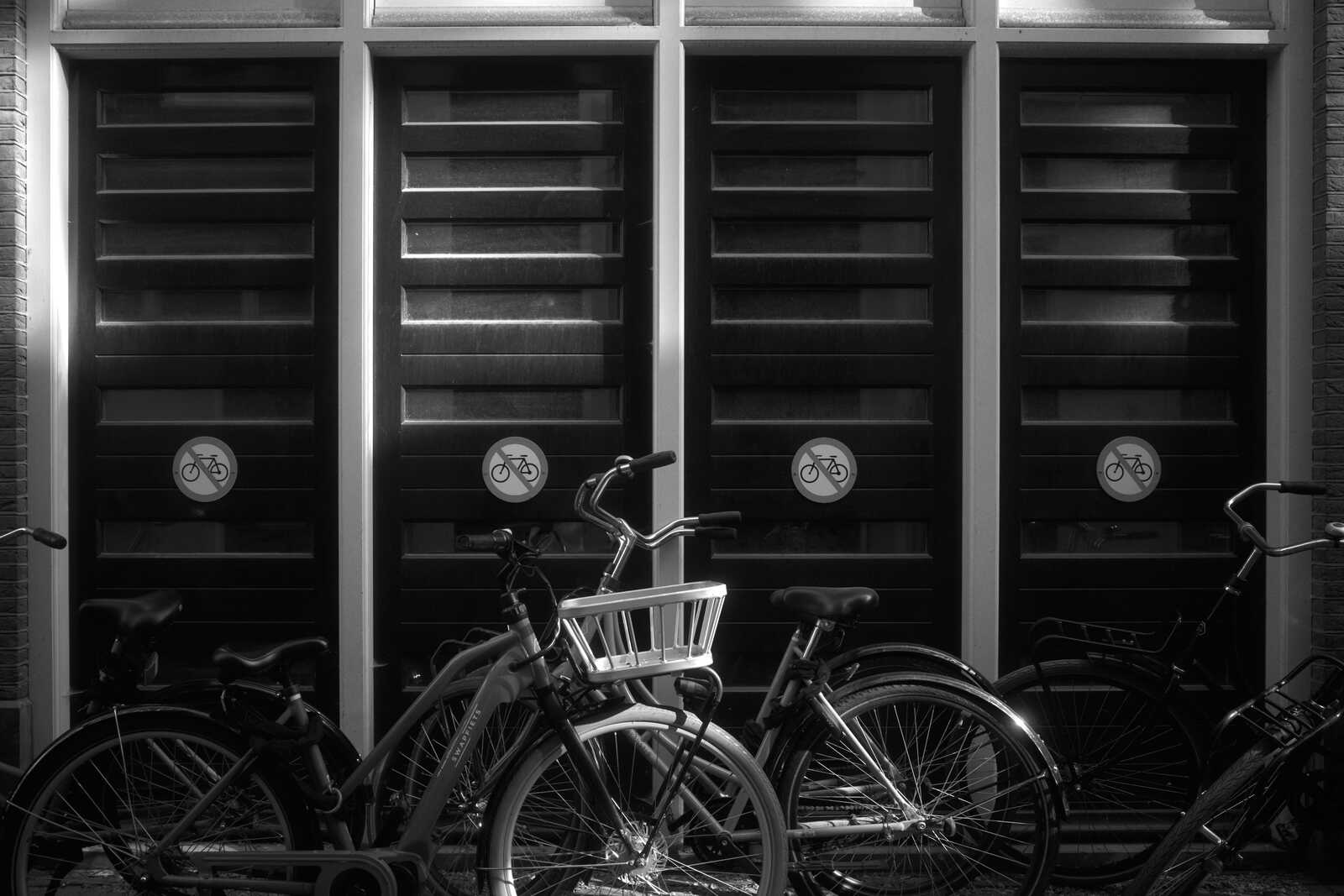 Three bicycles stand in front of a white door. Each bike has a basket on the handlebars. The door has three circular windows with bicycle symbols. The scene is in black and white.