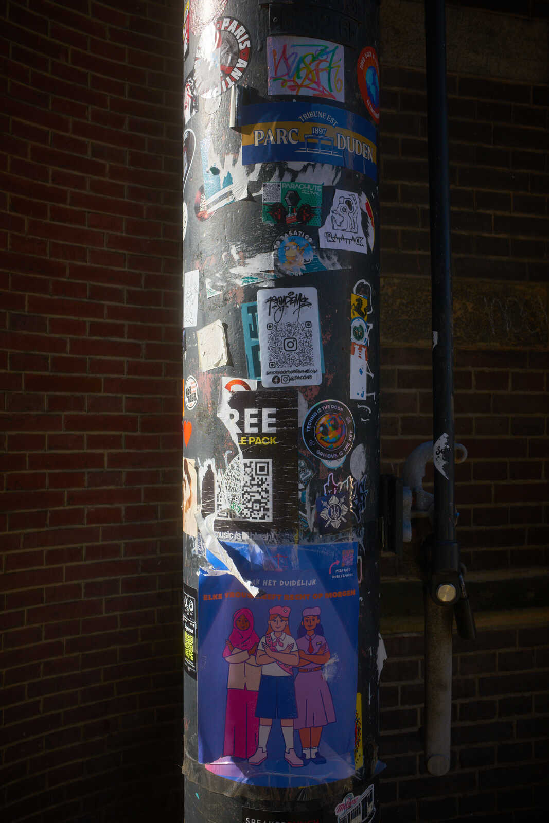 A metal pole covered in various colorful posters and stickers, set against a dark brick wall. Two figures are depicted on a poster, facing forward.
