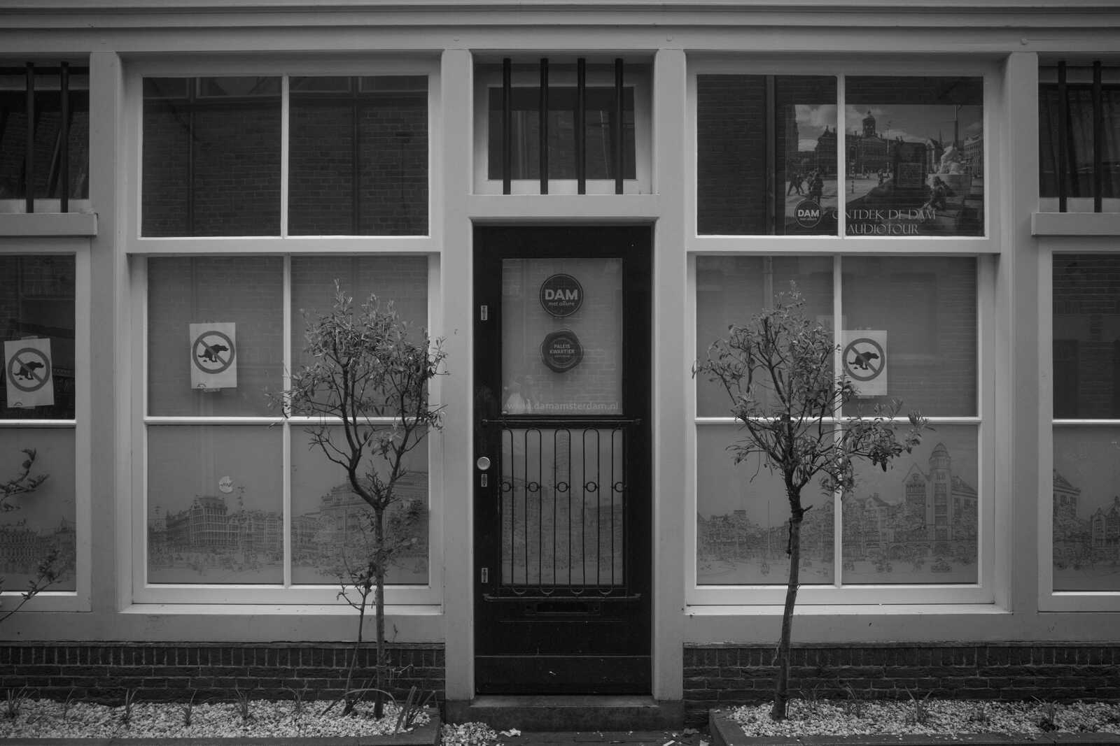 Black and white photo of a black door with a grille and a small window. Sign "DAM" is visible. Two small trees flank the door. A window is behind the door with a landscape displayed.