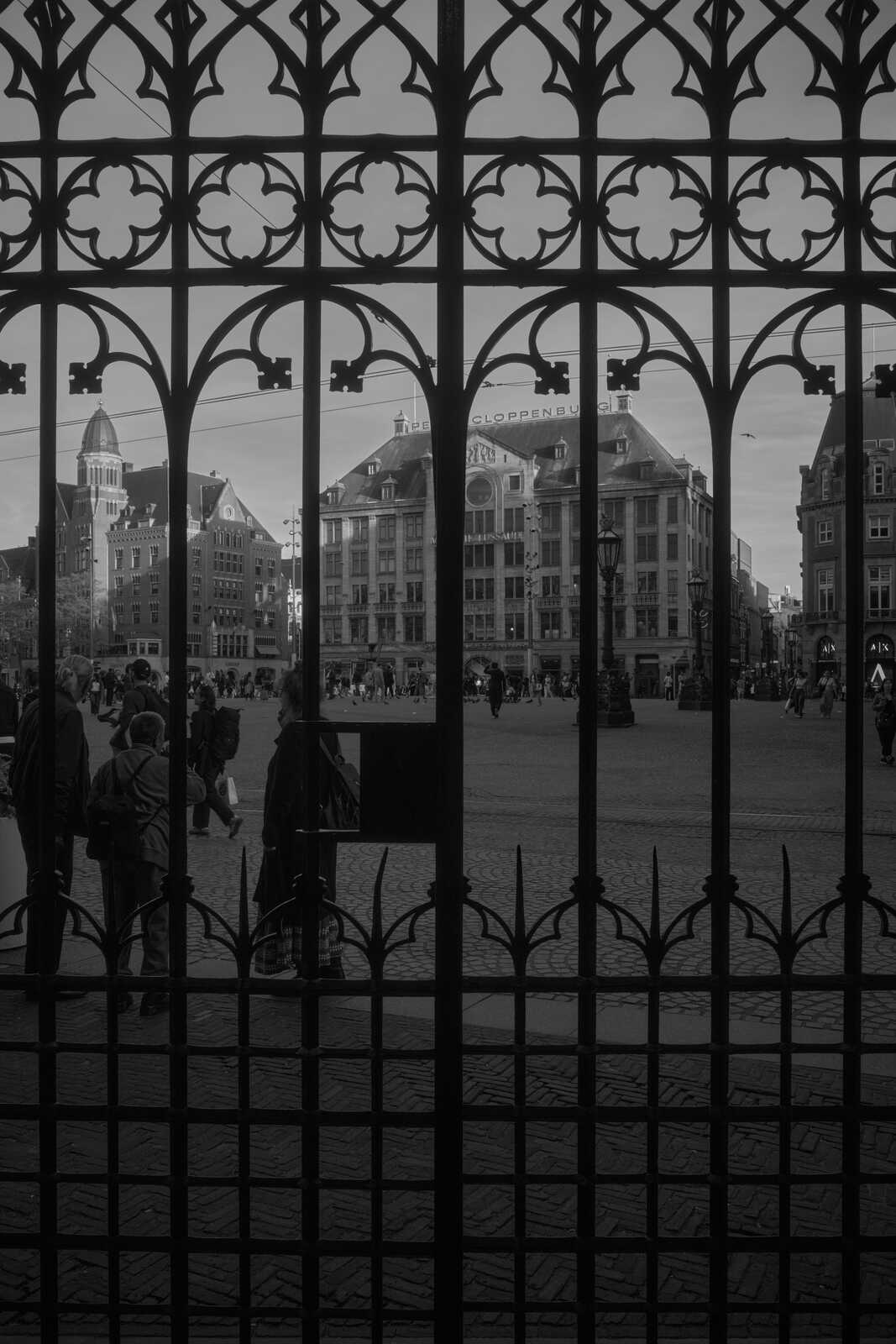A black and white photo of a city street viewed through a wrought-iron gate. Several people are walking on the street, and buildings are visible in the background. A sign reads "CLOPPENBURG".