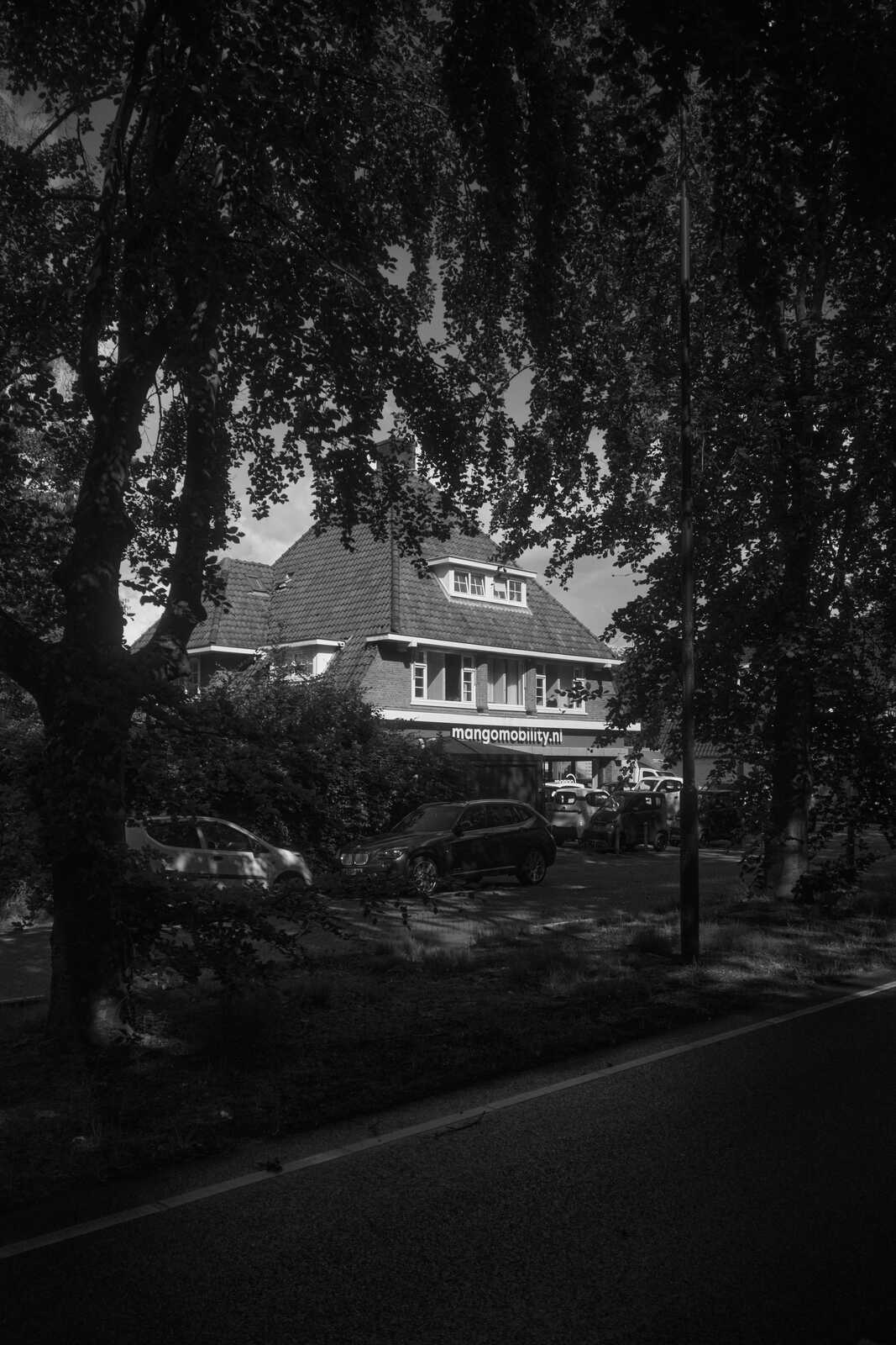 Black & white image of a building with "mangomobility.nl" signage, framed by trees and viewed from across a road. Cars are parked nearby.