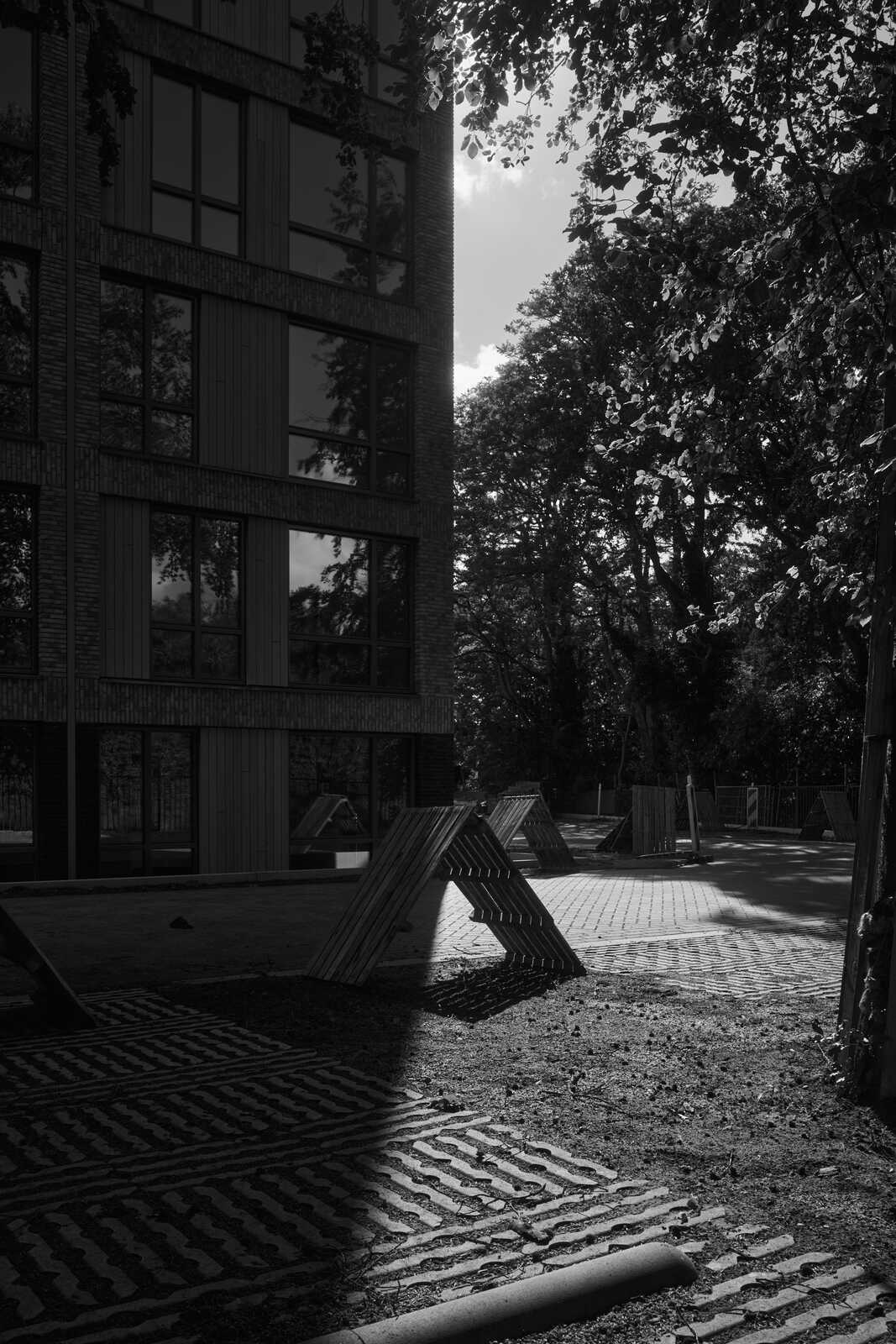 Black and white image showcasing a modern building facade beside trees and a playground with geometric climbing structure. Strong shadows are present.