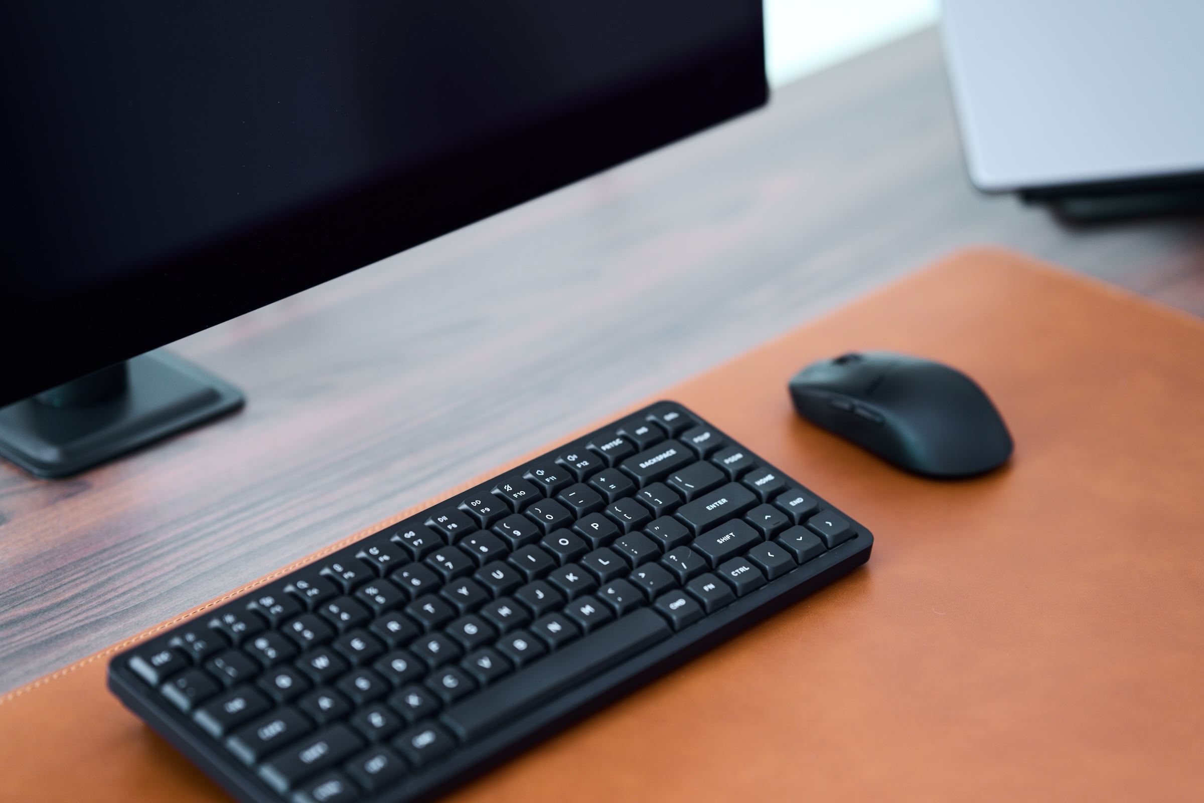 A black keyboard and mouse sit atop a brown desk mat in front of a computer monitor on a walnut desk.