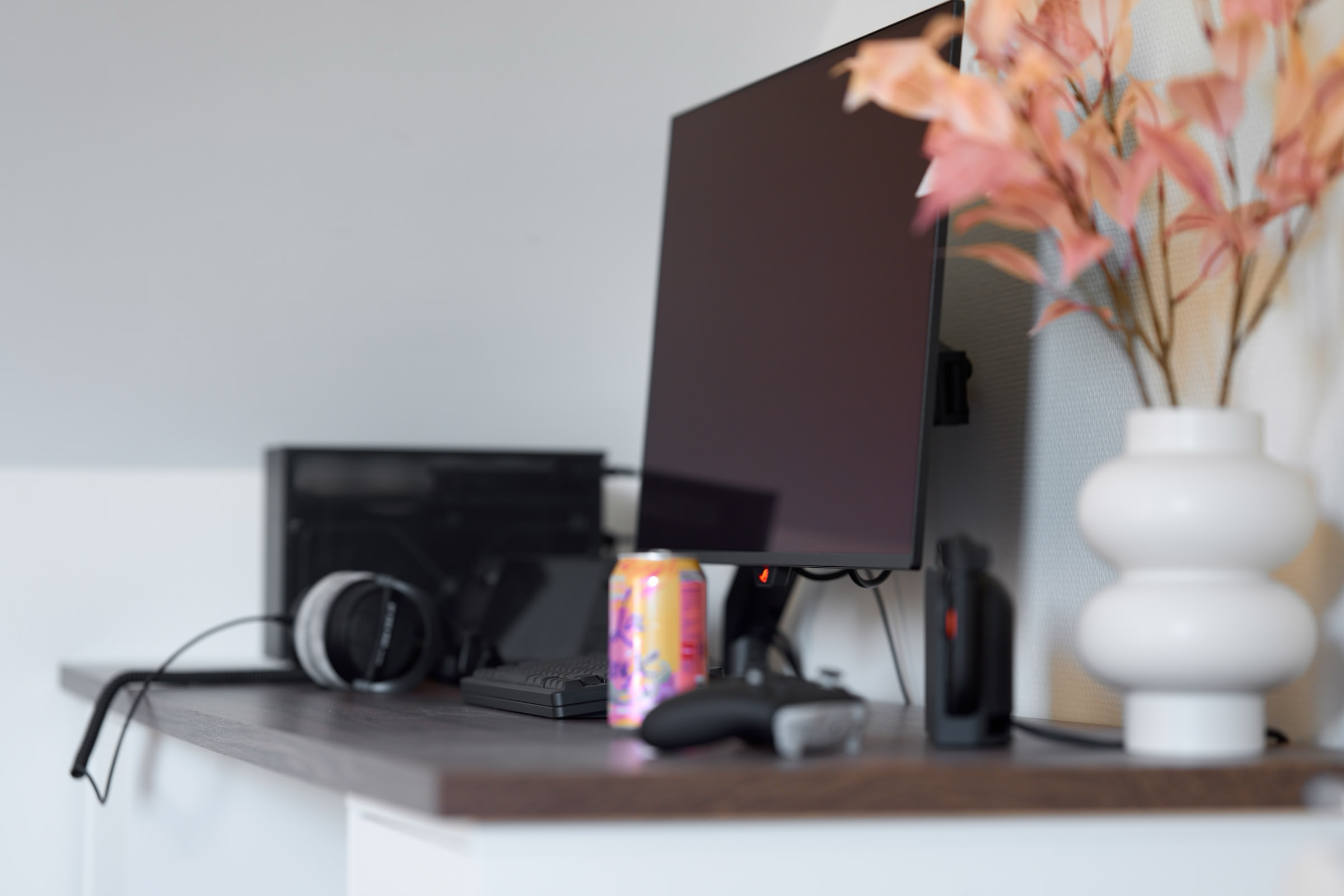 A modern desk with PC, a large monitor, keyboard, mouse, headphones, gamepad, Nintendo Switch 2, and a soda can. A vase with pink flowers sits to the right.