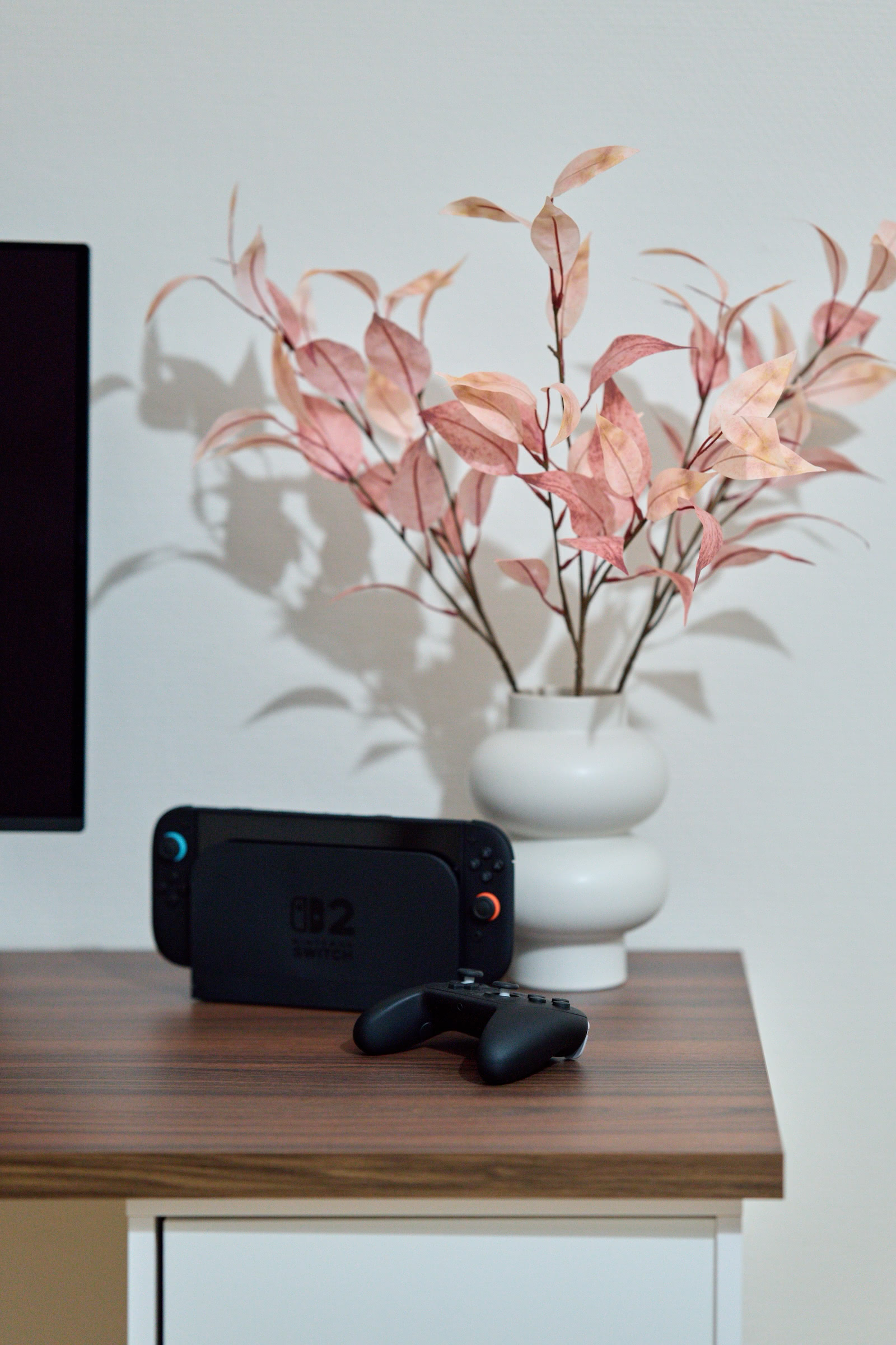 Nintendo Switch 2 with Pro Controller sits atop a walnut desk. A vase holding pink branches is behind it, casting shadows on the wall.