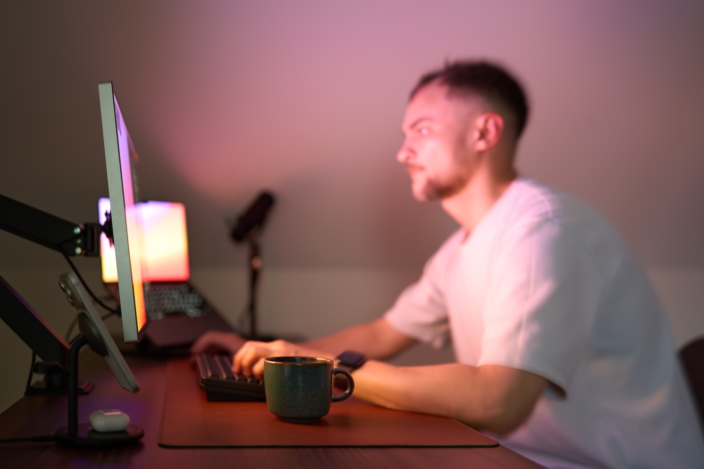 A person sits at a desk in front of a monitor. He's typing, with a coffee cup and mouse nearby. Soft pink light coming from the display illuminates the scene.