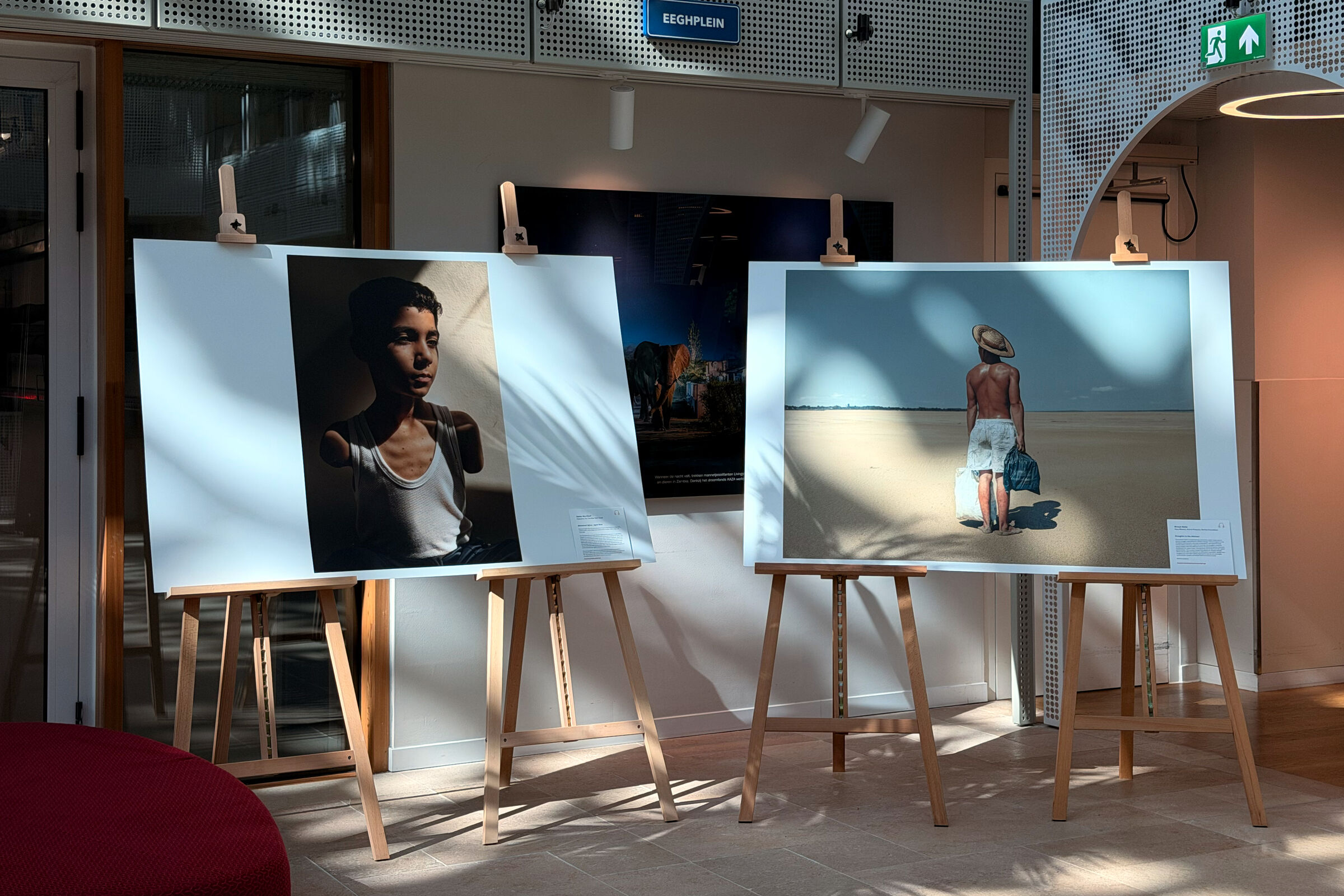 Two framed photographs from World Press Photo displayed on wooden easels stands. One depicts a boy with amputaded arms, the other a landscape with a distant figure. Both are under soft lighting.