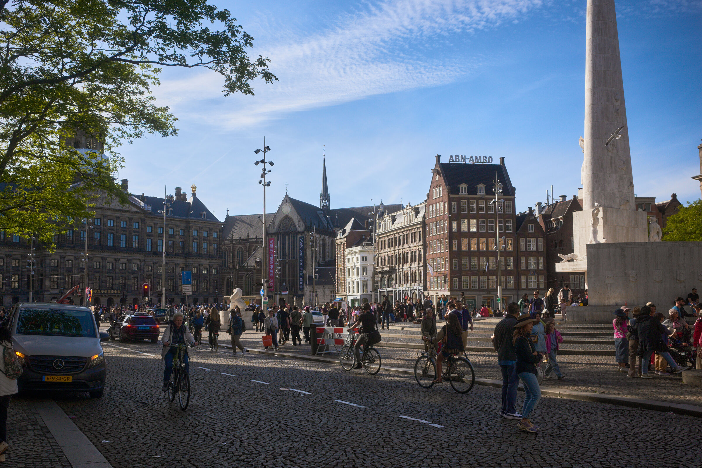 Dam square in Amsterdam. A cobblestone street with people and bikes. Ornate buildings with gabled roofs line the street. De Nieue Kerk is visible in the background under a blue sky.