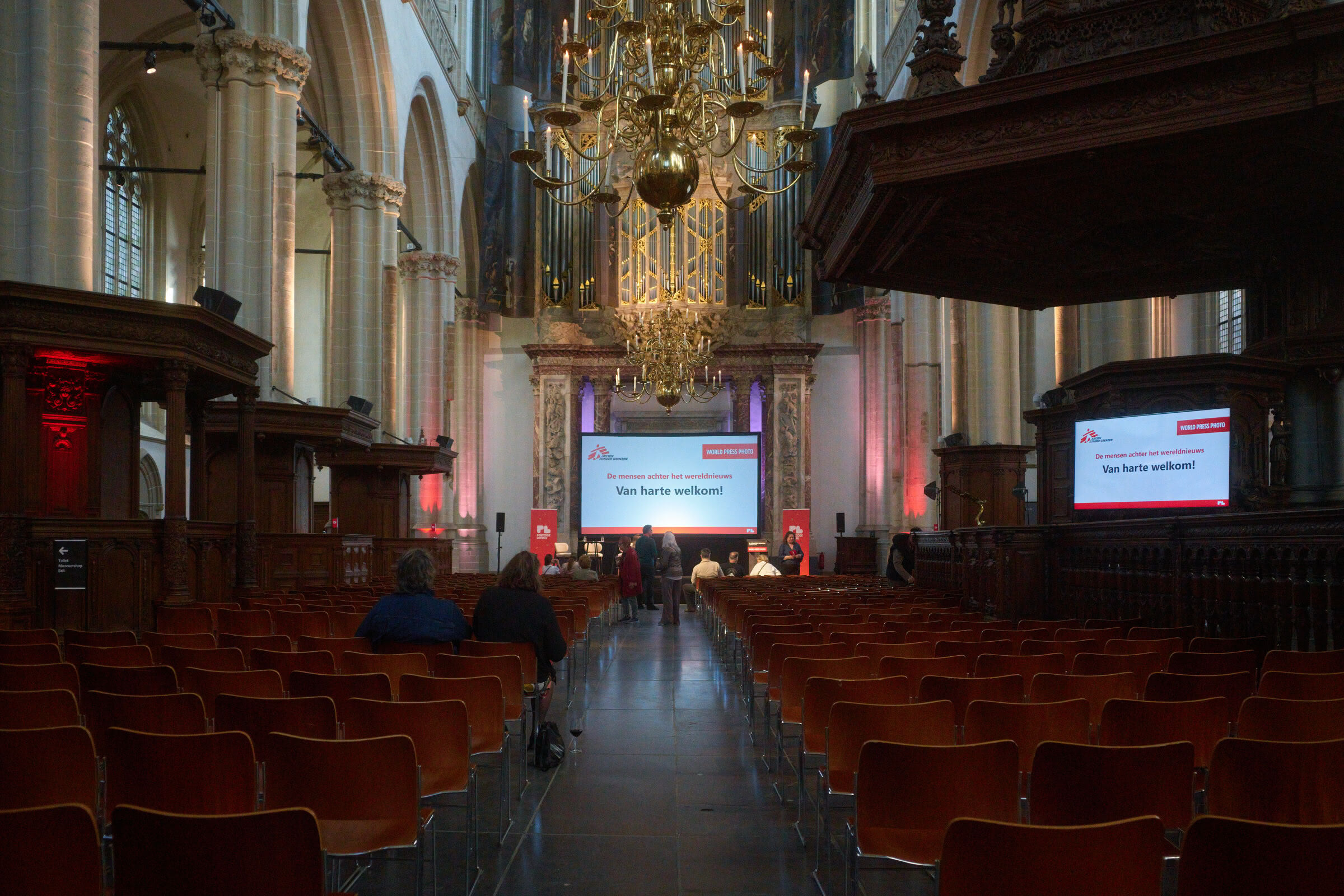 An expansive interior with rows of red seating facing a raised stage. Pillars, ornate details, and screens with text are visible. A chandelier hangs above the stage.