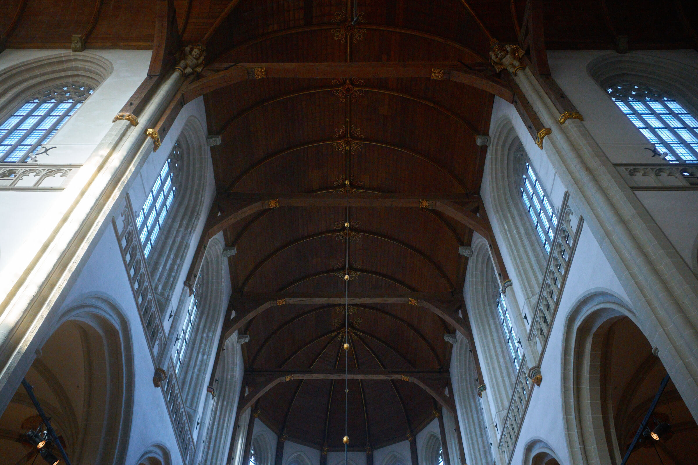 A cathedral interior view. Vaulted wooden ceiling with beams and a chandelier. Stone pillars and arched windows with stained glass are visible on either side.