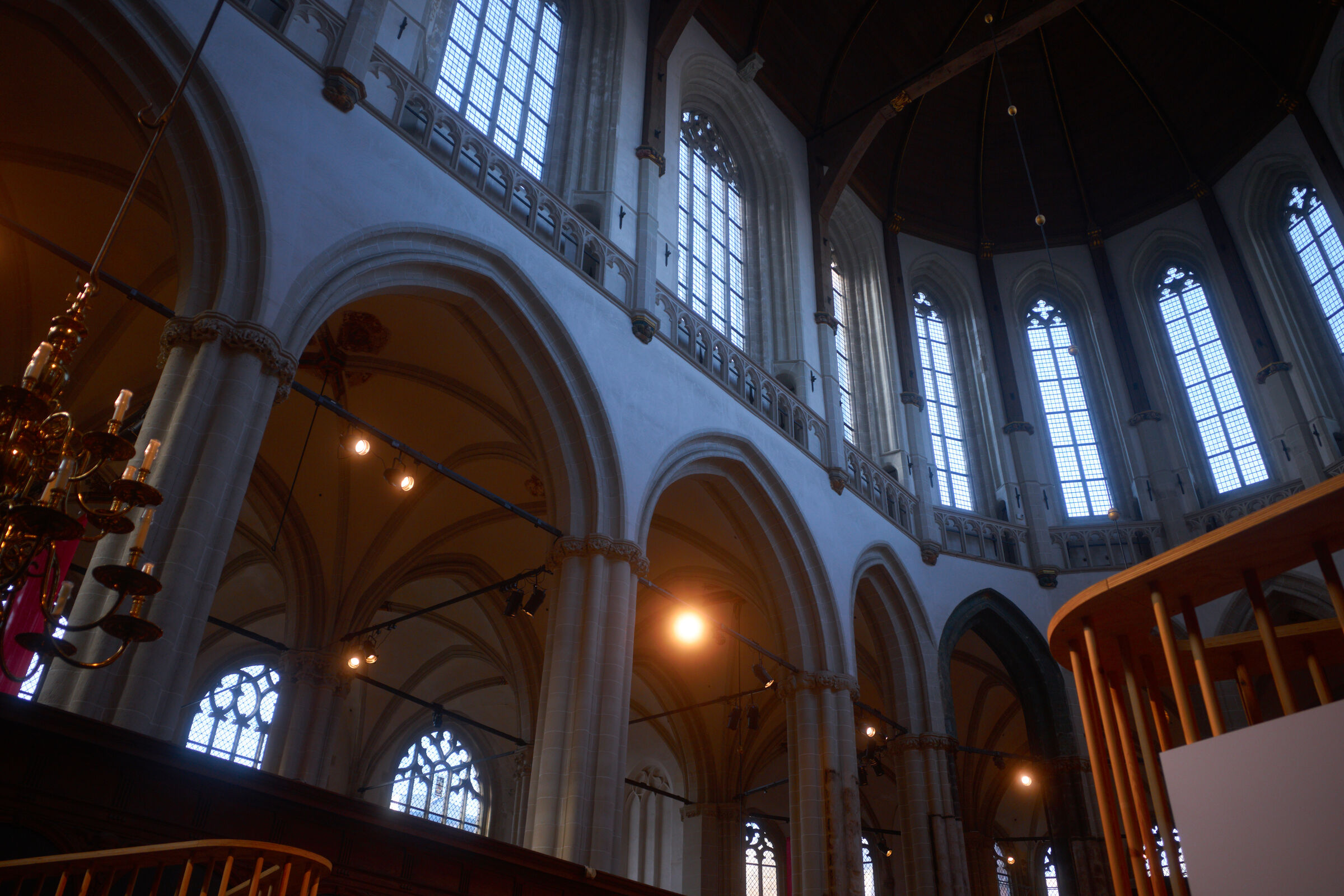 Tall arched windows and stone pillars create a grand interior. Balconies with wooden railings and a chandelier are visible. Light streams through the windows.