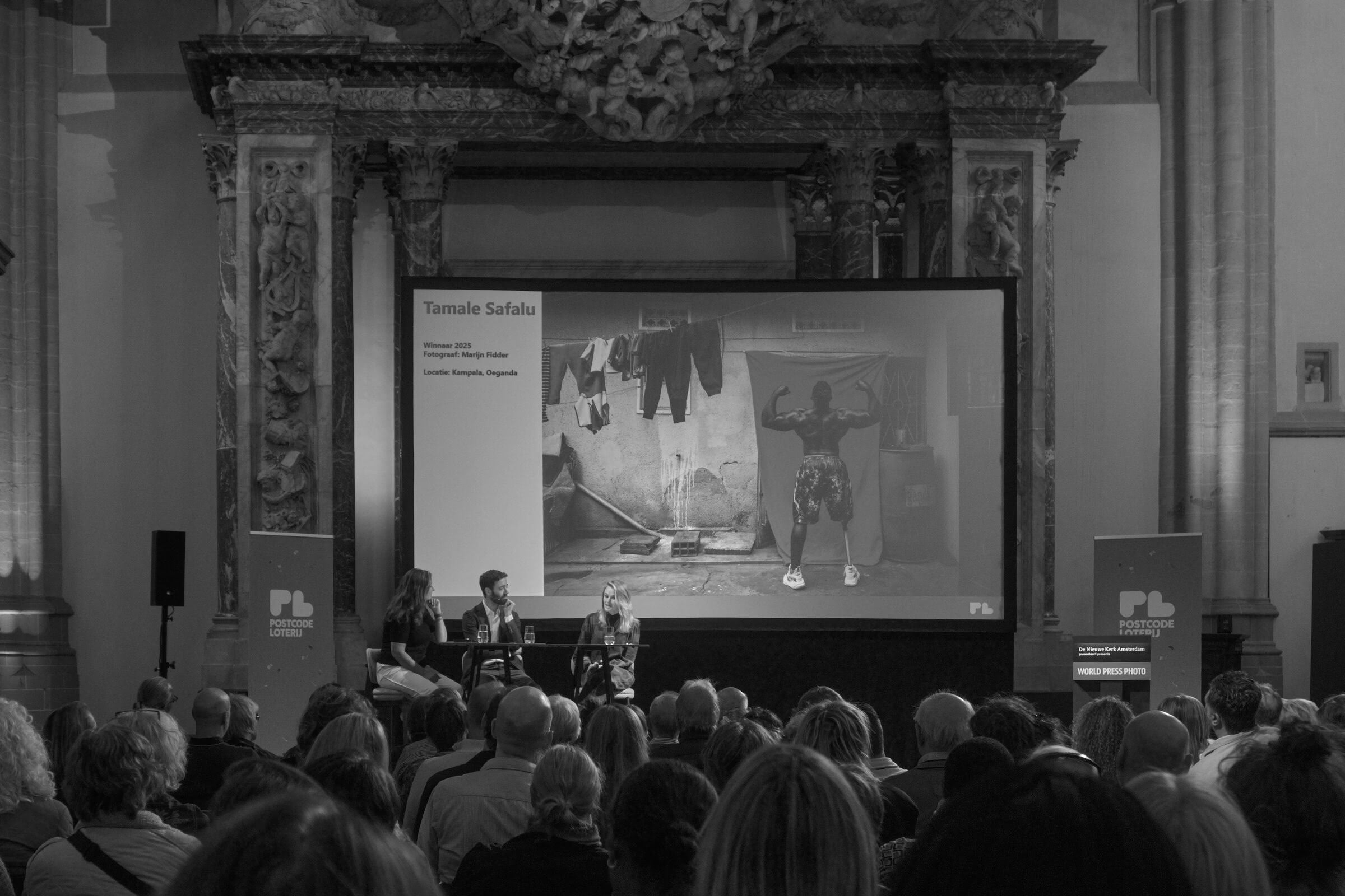 A picture of a man without one leg, posing, and flexing muscles is displayed on a screen, next to a text: Tamale Safalu. An audience sits in rows behind him. Two figures are visible at the front of the room.