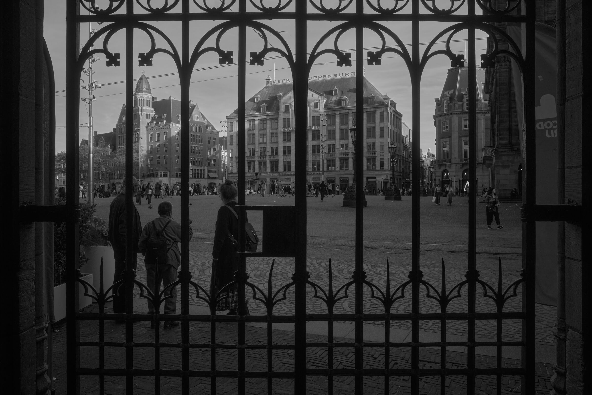 Figures stand before an ornate gate overlooking Dam square with buildings and people. A large tower rises in the background. The scene is in black and white.